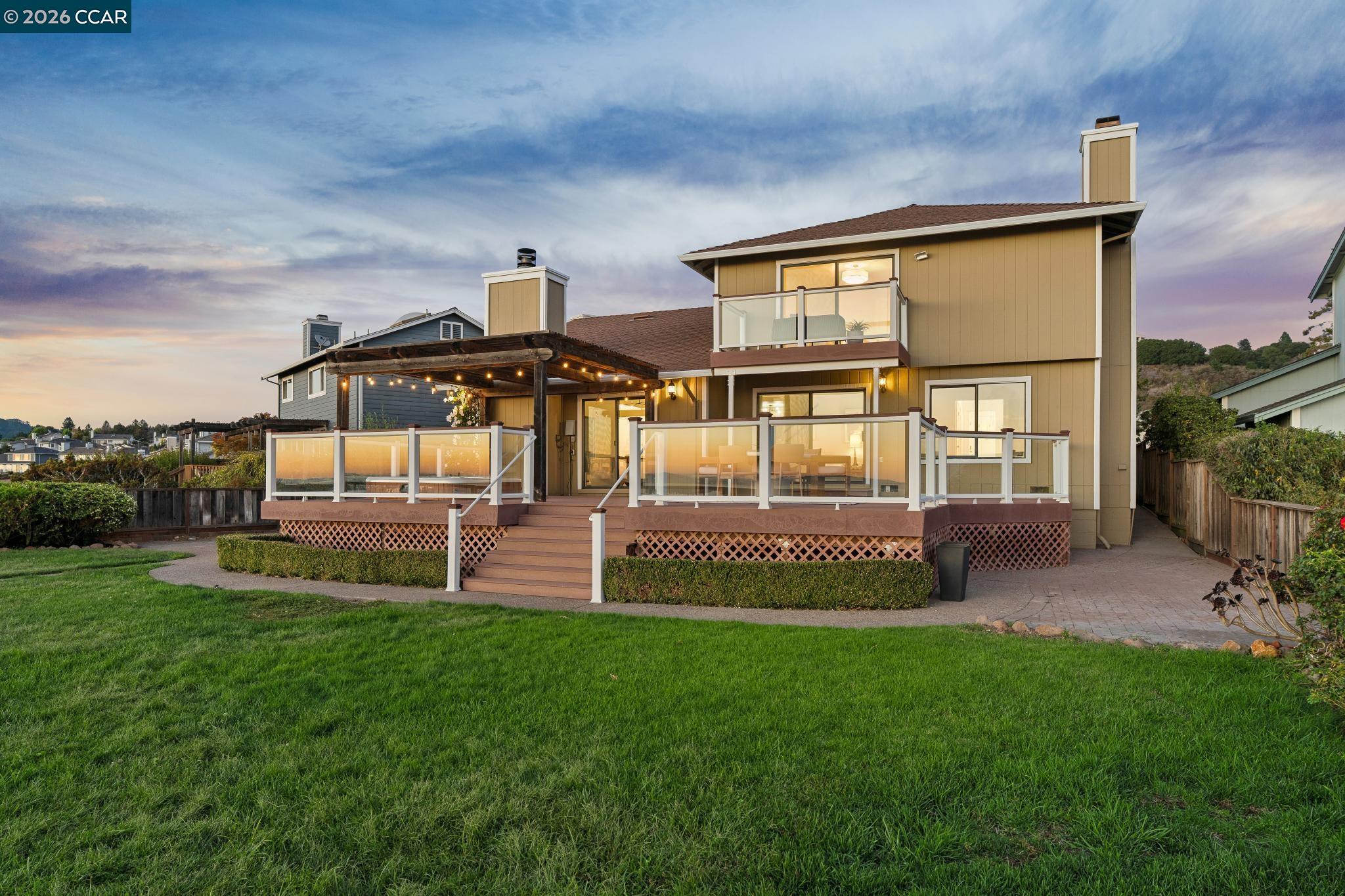Two-story tan house with a glass-railed deck, pergola lights, and a broad lawn at sunset.