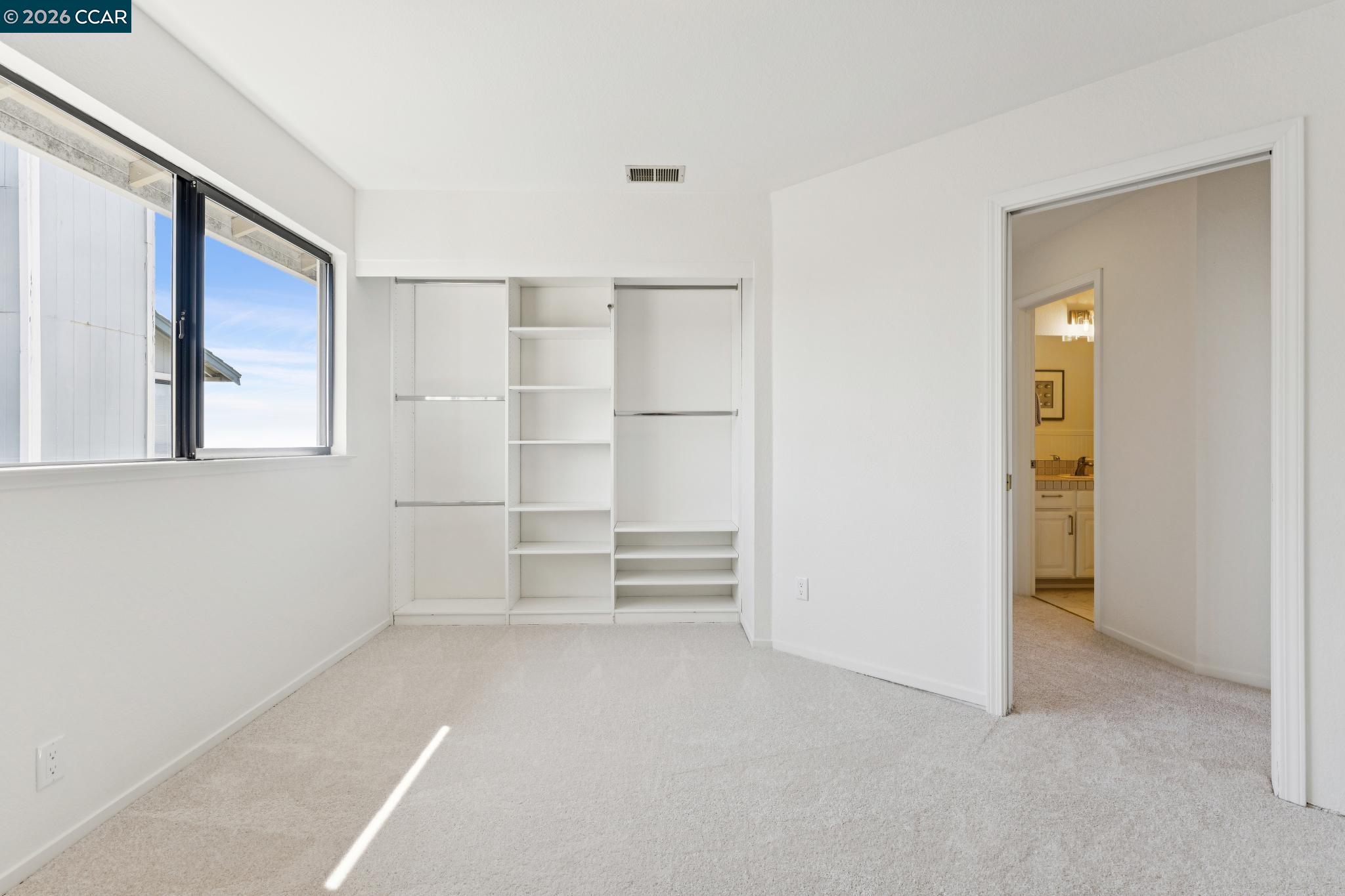 Vacant bedroom with built-in closet shelving, carpet, and a window beside the bathroom doorway.