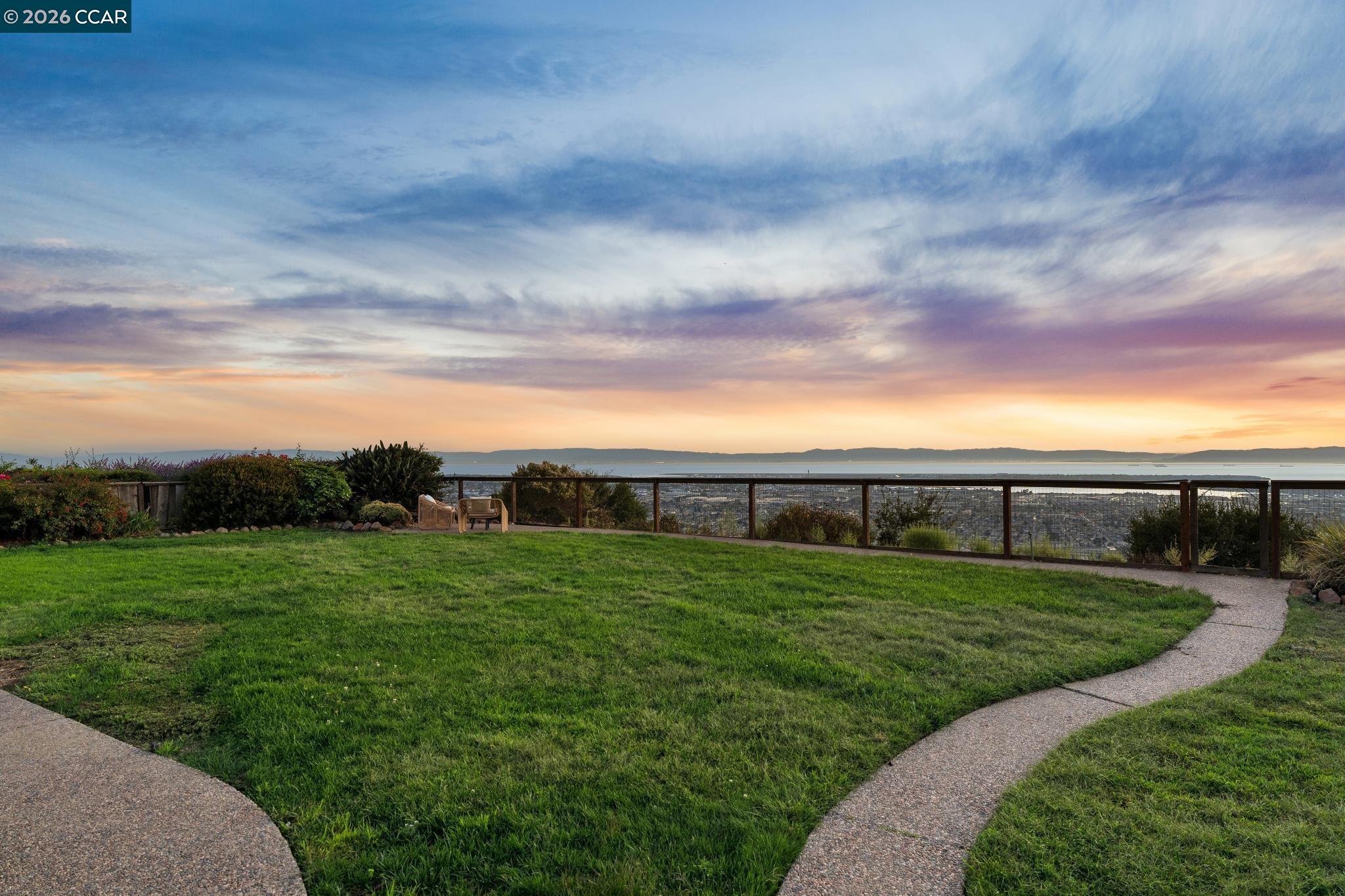 Backyard lawn and curved paths overlook the water and distant city beneath a colorful sunset sky.
