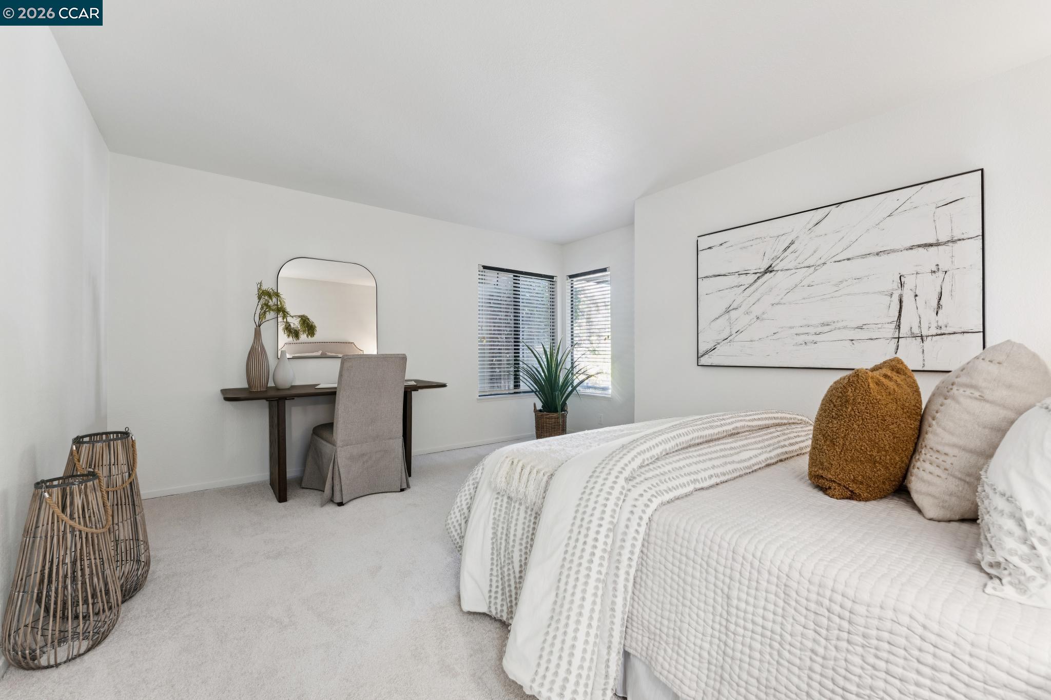 Staged bedroom with a white quilted bed, dark writing desk, woven baskets, and large abstract wall art.