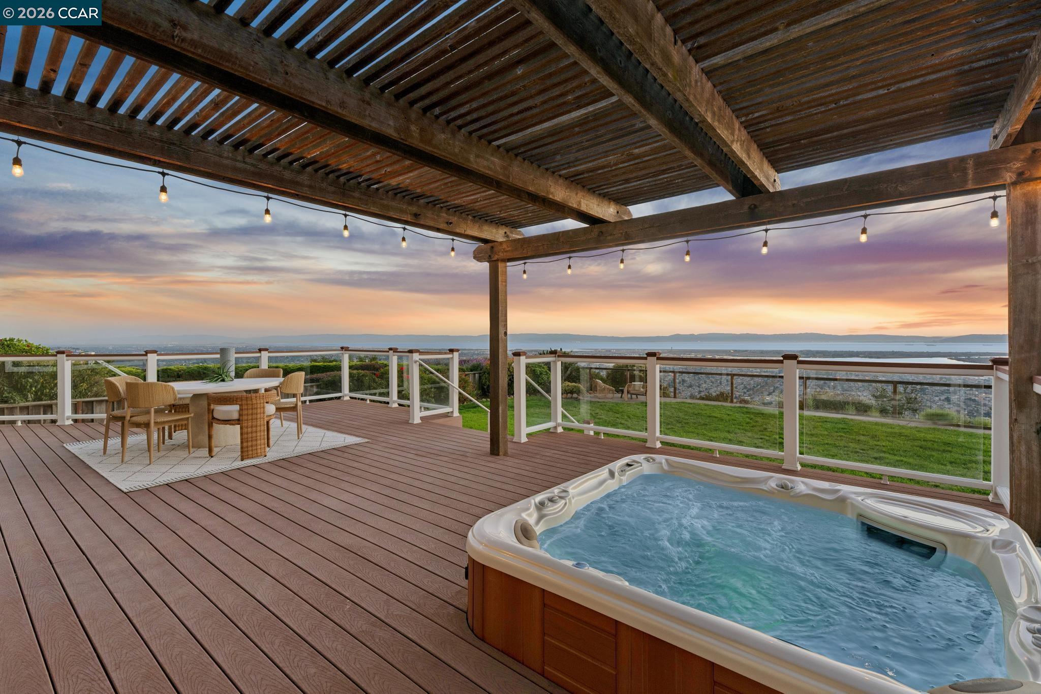 Deck hot tub and dining area beneath a timber pergola, overlooking water at sunset.
