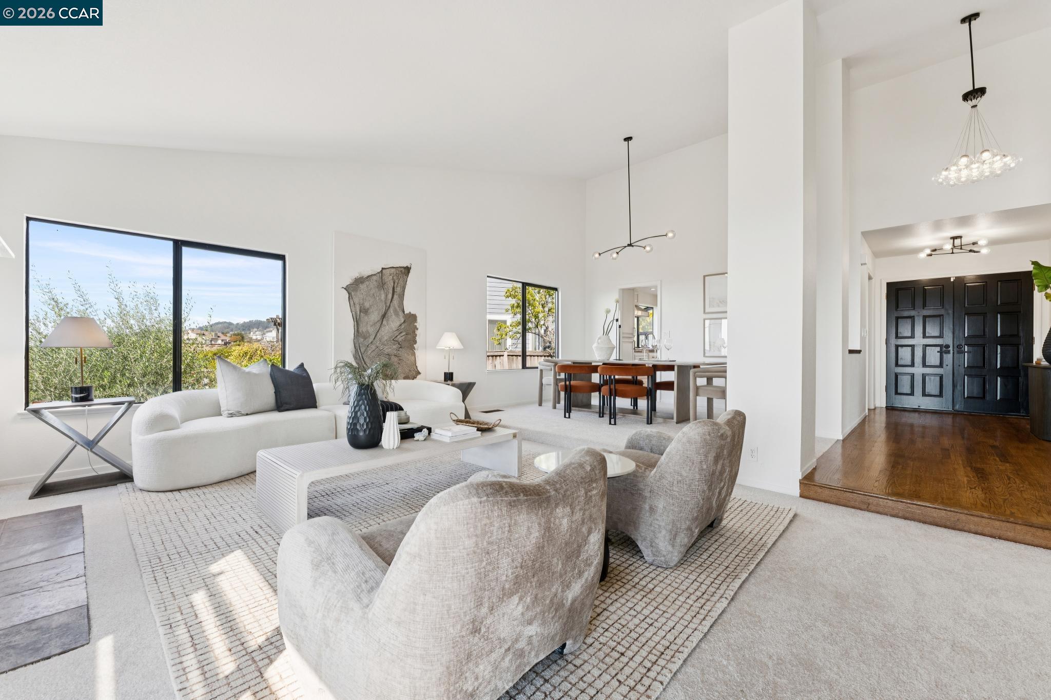 Open-plan living and dining room with high ceilings, large windows, and black double entry doors.