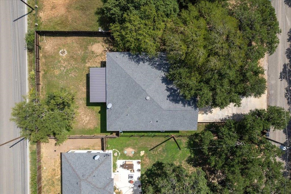 Aerial view of a gray-roofed home, fenced yard, mature trees, and patio beside the road.