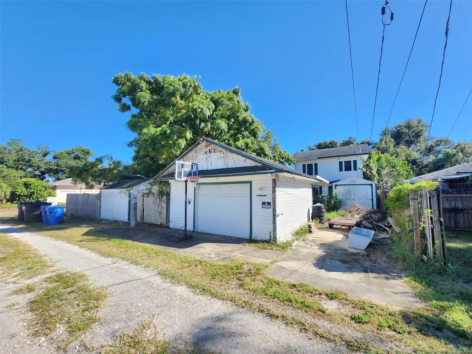 Weathered white detached garage with green trim, a basketball hoop, and a cluttered driveway.