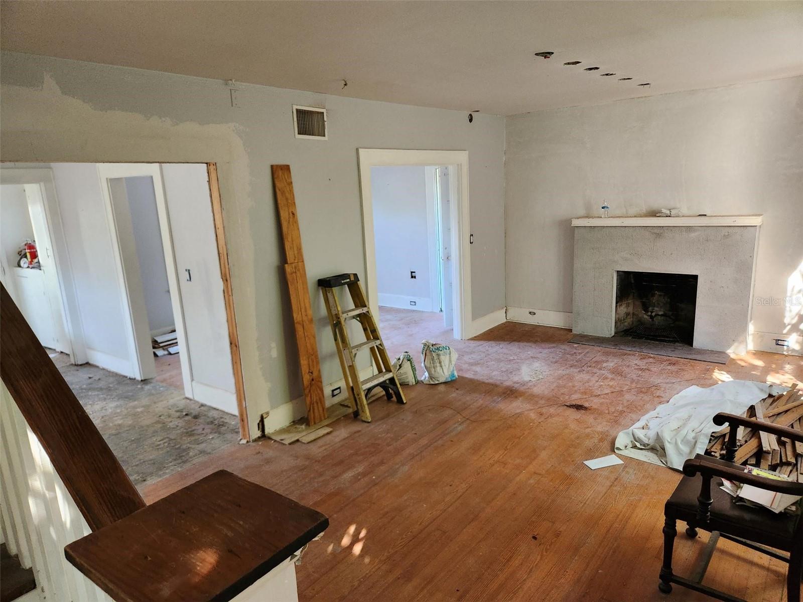 Living room under renovation with a fireplace, worn wood floors, and construction materials.