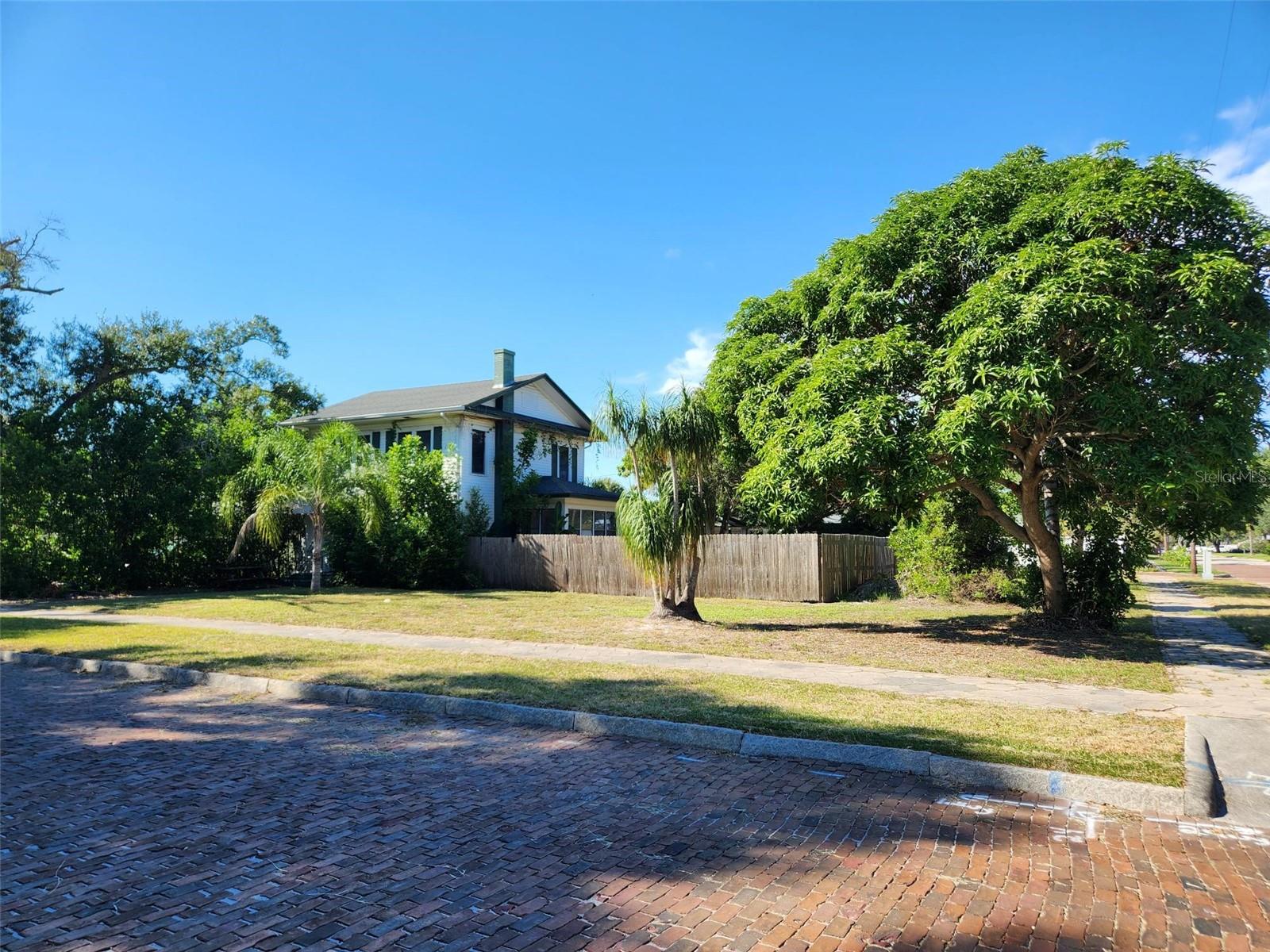 Two-story house behind a wooden fence, framed by palm trees and a large shade tree.