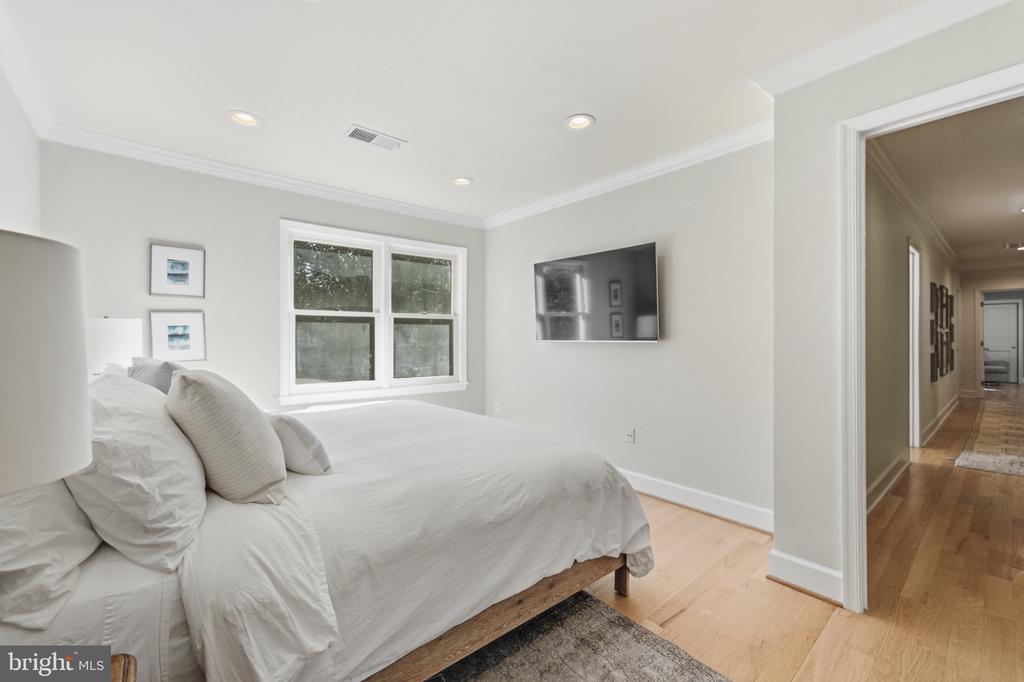 Bedroom with a white bed, large window, wall-mounted television, and doorway to a hardwood hallway.