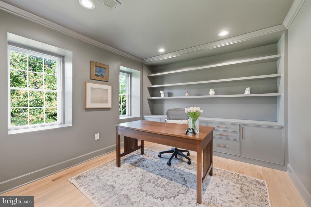 Home office with built-in gray shelving, a wooden desk, and windows overlooking greenery.