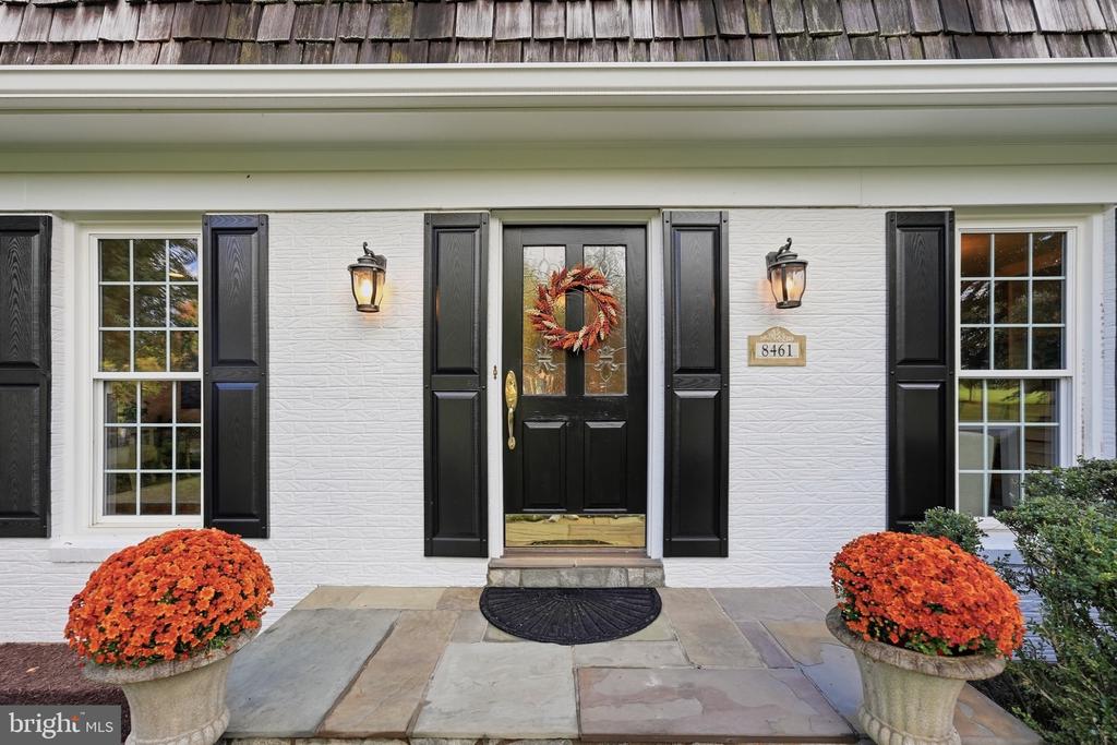 White-brick home entrance with black shutters, a wreath-adorned door, stone walkway, and orange flower planters.