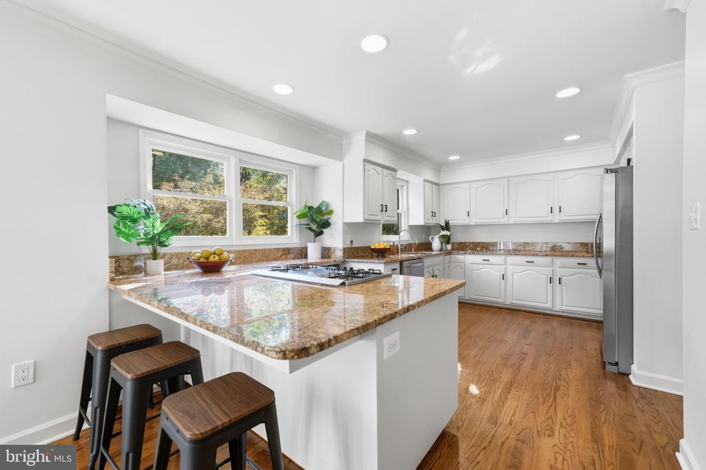 Kitchen with a granite island and stools, white cabinetry, hardwood floors, and windows overlooking trees.