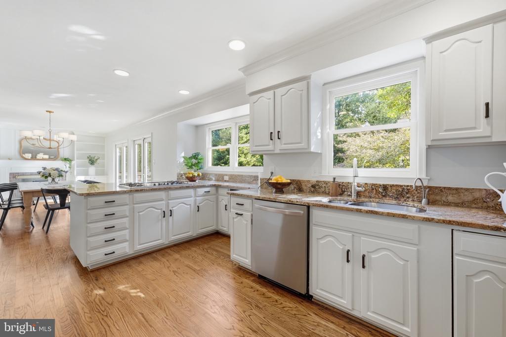 Bright white kitchen with granite counters, stainless dishwasher, hardwood floors, and windows overlooking trees.