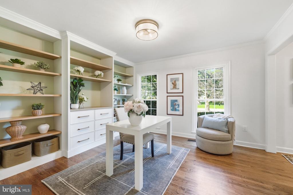 Staged home office with built-in shelves, a white desk, and two windows overlooking greenery.