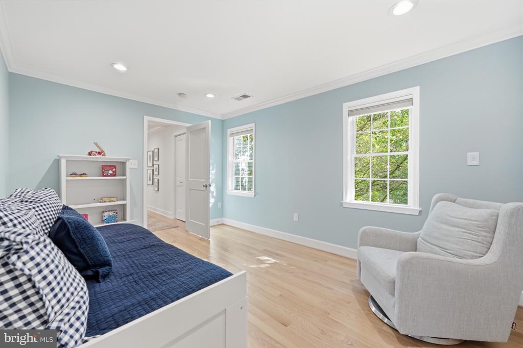 Bedroom with pale blue walls, navy bedding, a gray armchair, and windows overlooking leafy trees.