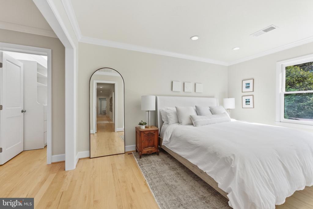 Bedroom with a large white bed, arched mirror, light wood floors, and a window overlooking greenery.
