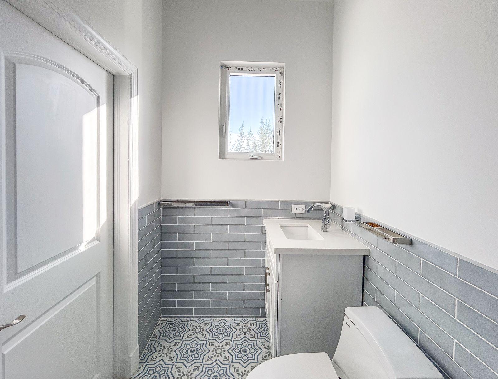 Bathroom with gray tile walls, patterned floor, white sink vanity, toilet, and a small window.