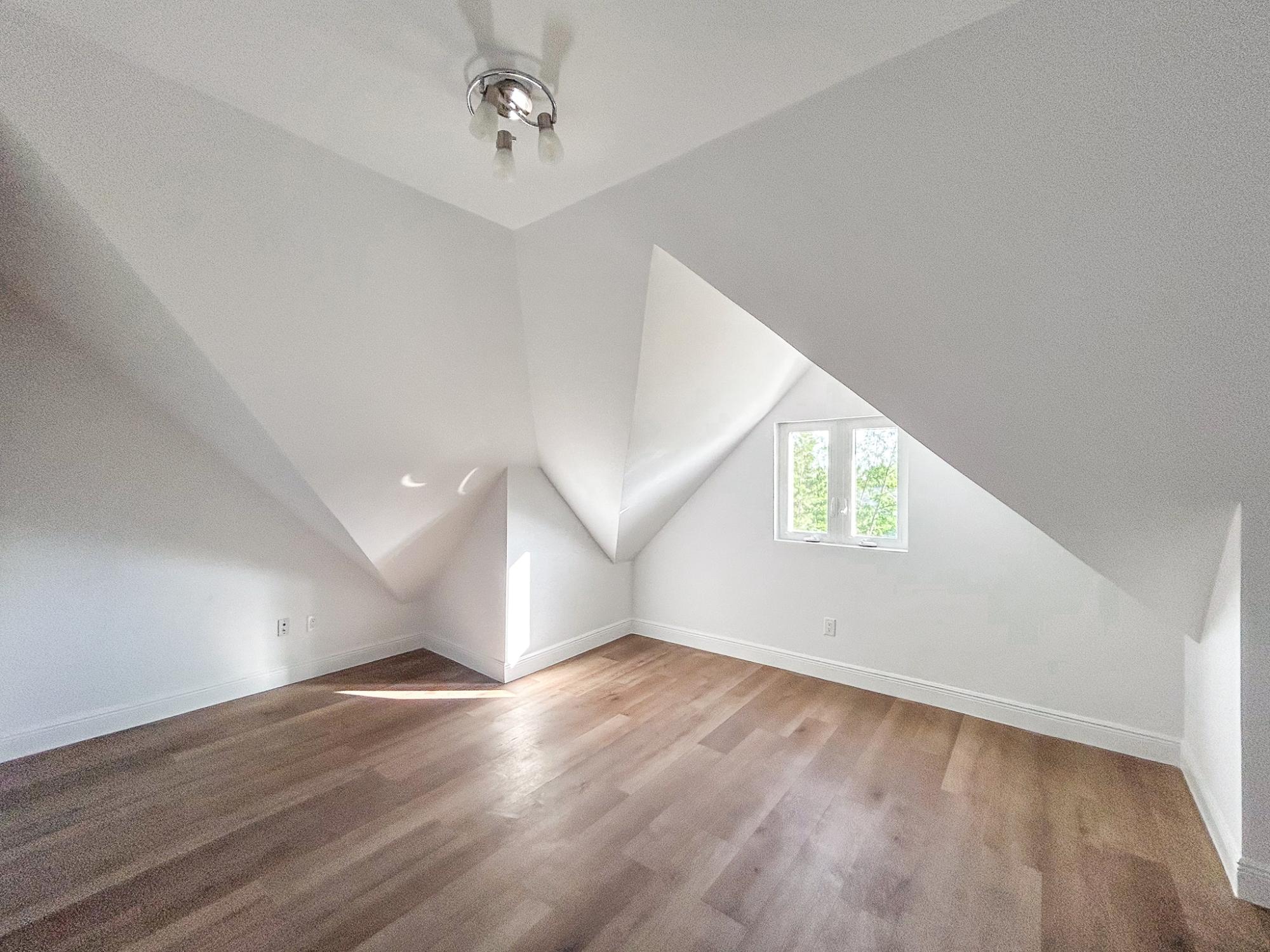 Empty attic room with sloped ceilings, light wood floors, and a small window