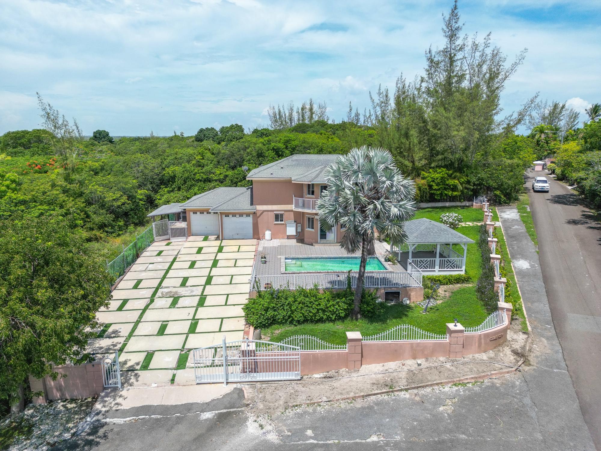 Two-story house with a pool, covered gazebo, and gated driveway amid tropical landscaping.