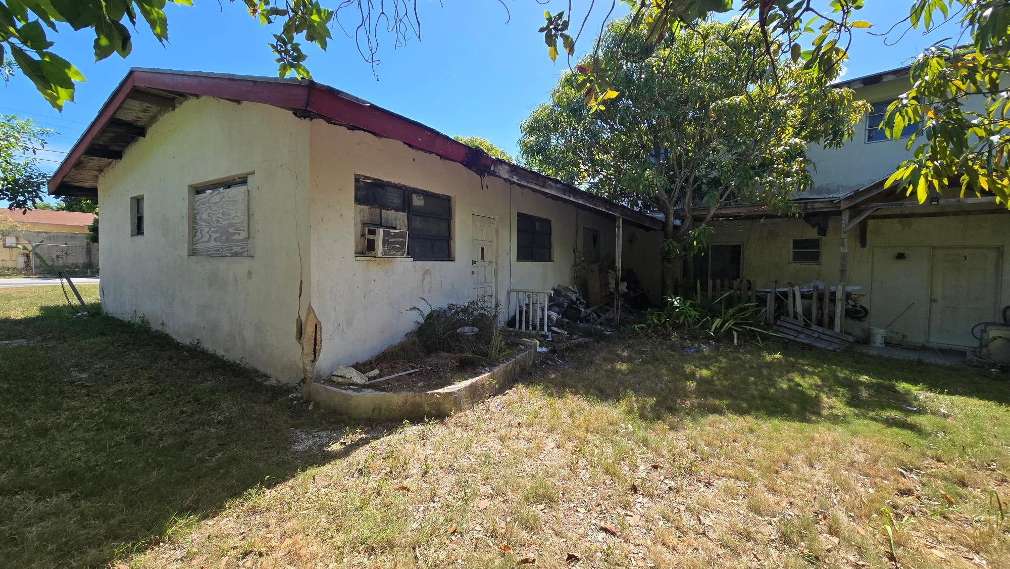 Weathered residential building with cracked walls, a boarded window, and debris beneath a shade tree.
