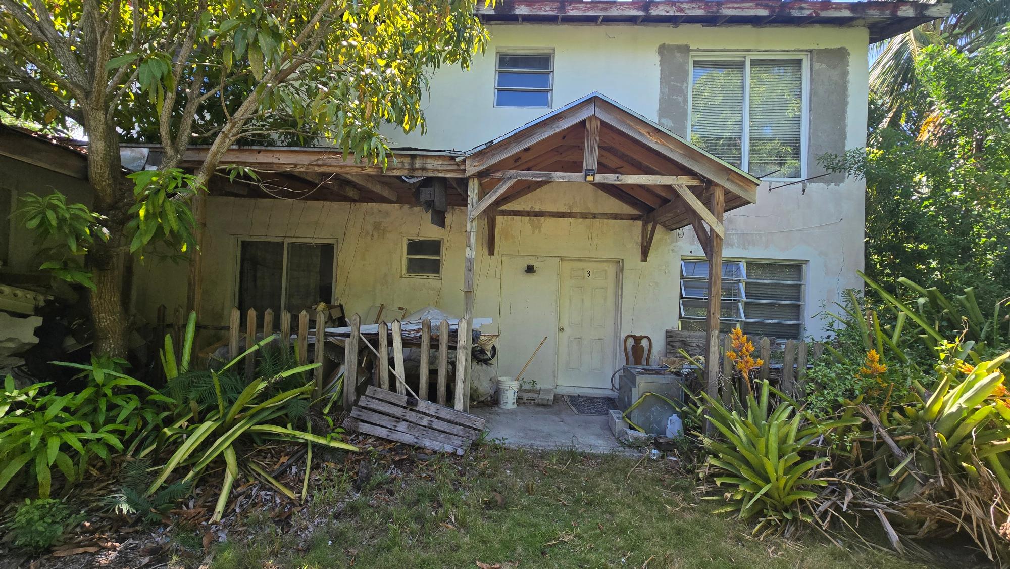 Weathered two-story house with a timber entrance canopy, small porch, and dense tropical foliage.