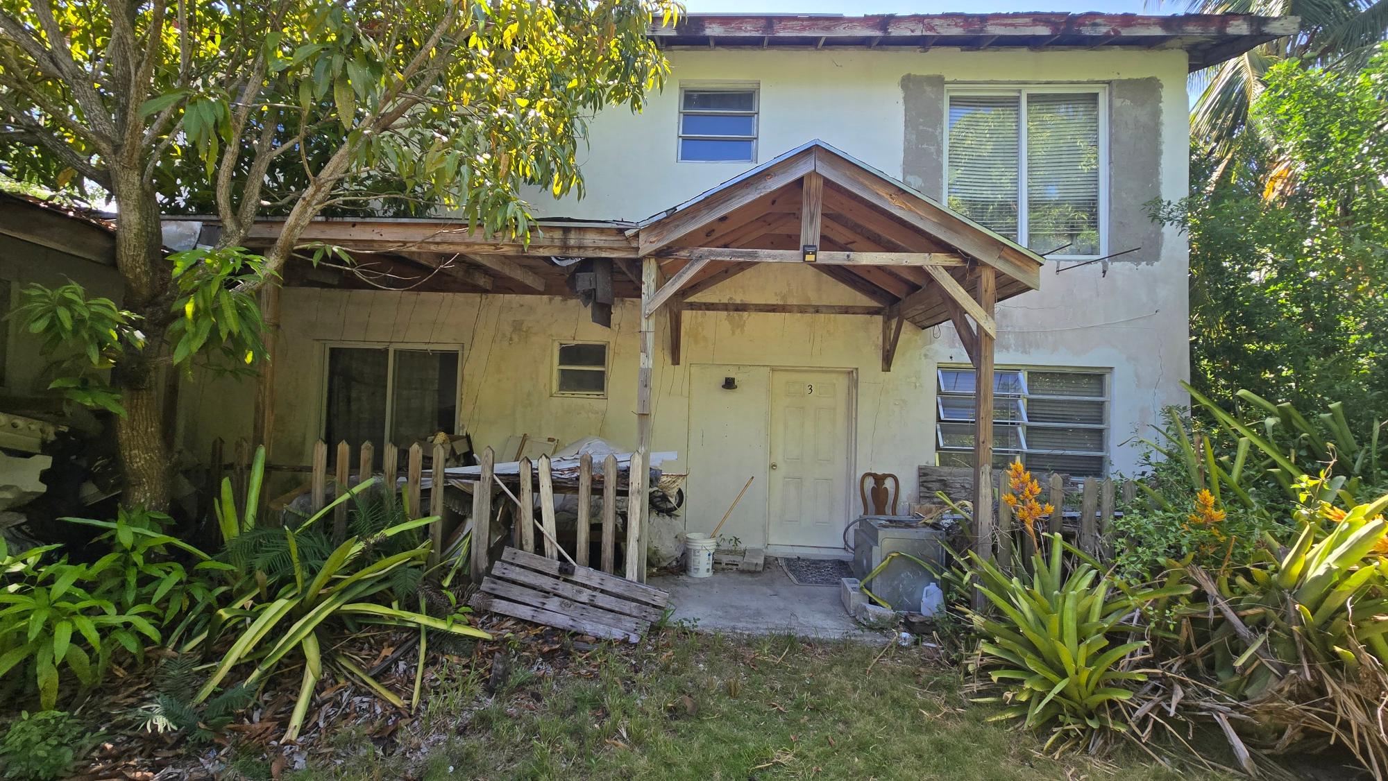 Weathered two-story house with a timber entrance canopy, surrounded by tropical plants.