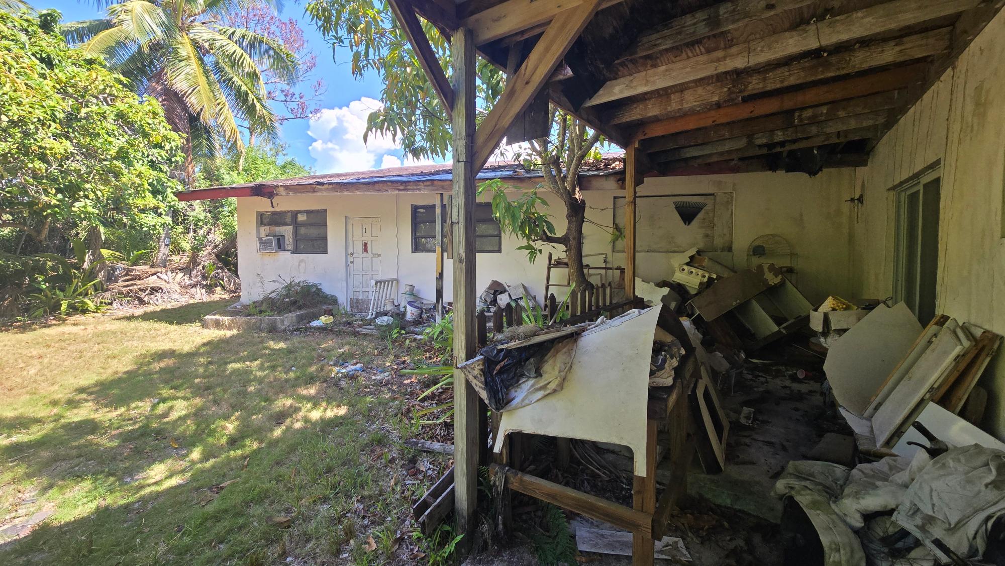 Weathered single-story building and debris-filled covered porch amid tropical greenery.