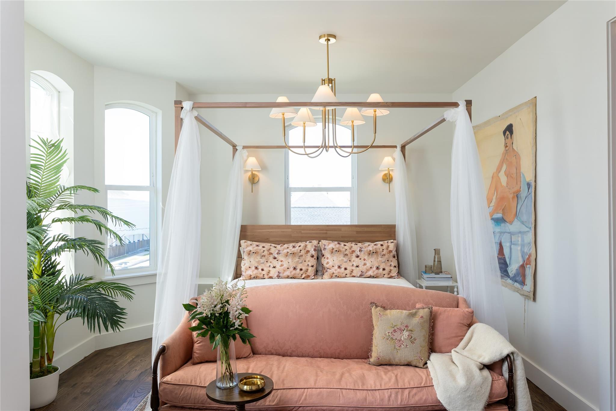 Bright bedroom with a sheer-draped canopy bed, tall arched windows, and a pink sofa at its foot.