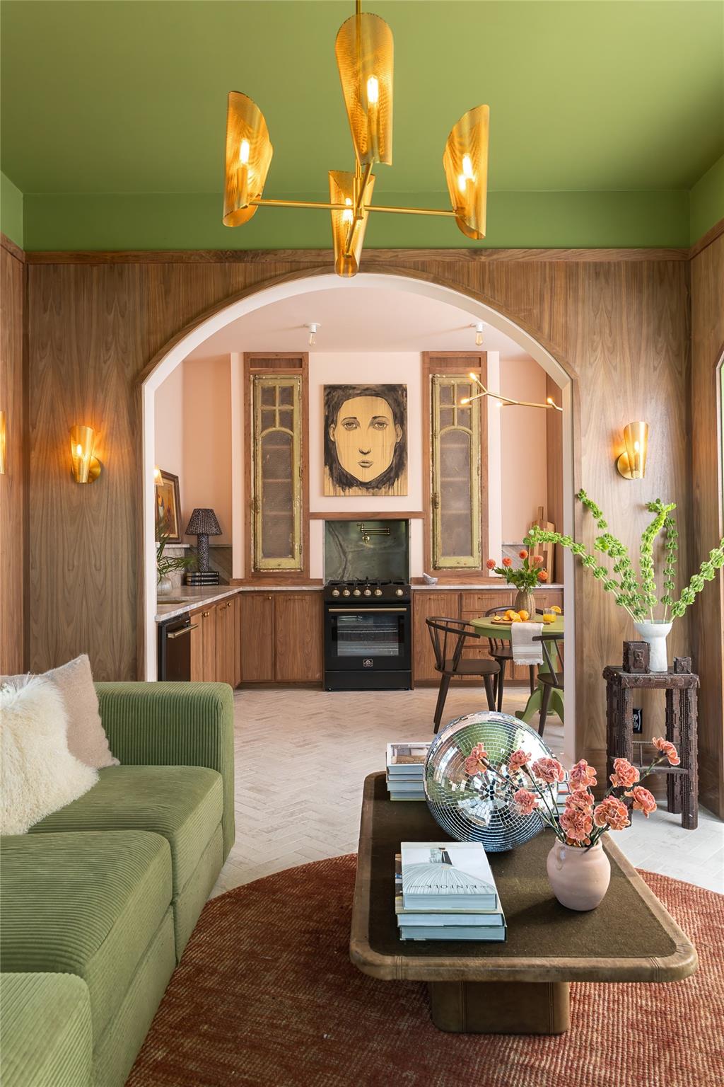 Furnished living room opening to a wood-paneled kitchen beneath a large arch and sculptural gold chandelier.