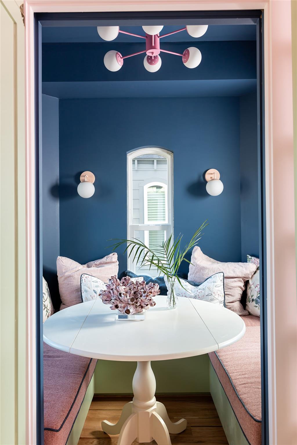 Dining nook with a white round table, pink banquette seating, and a pink chandelier against blue walls.