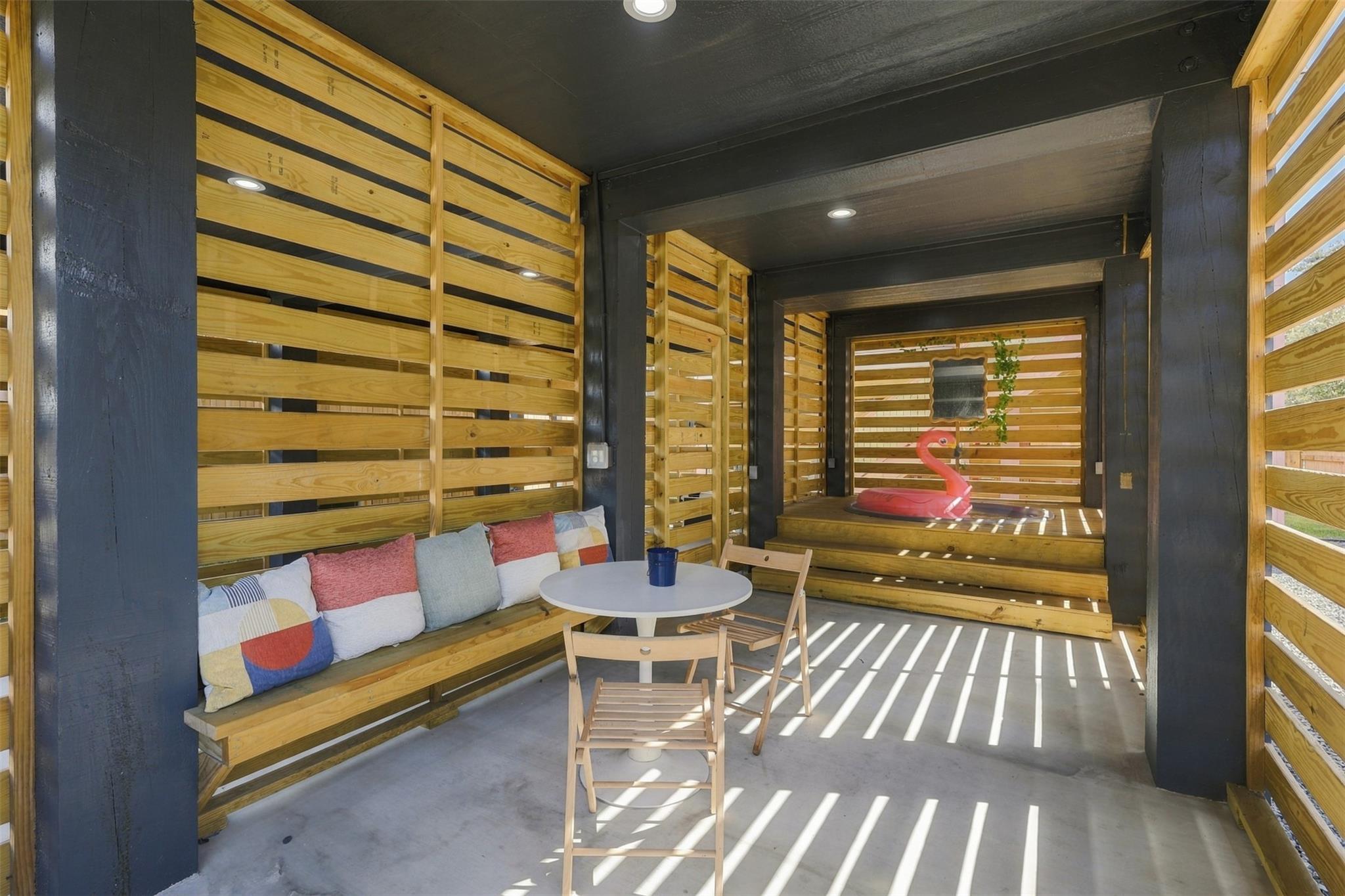 Covered patio with slatted wood walls, a cushioned bench, table and chairs, and a pink flamingo float.