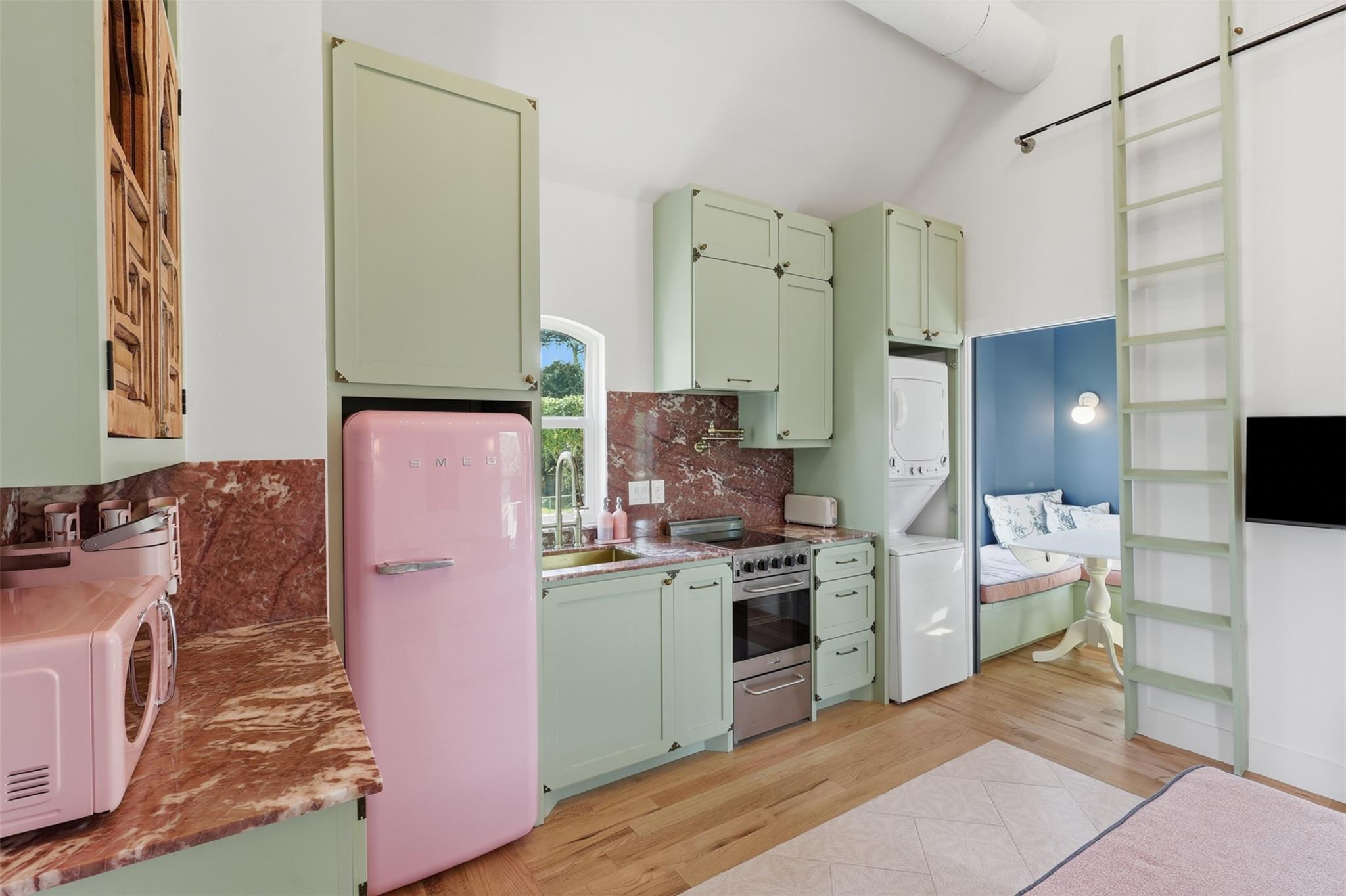 Kitchen with pale green cabinets, red marble counters, and a pink SMEG refrigerator beside a small sleeping nook.