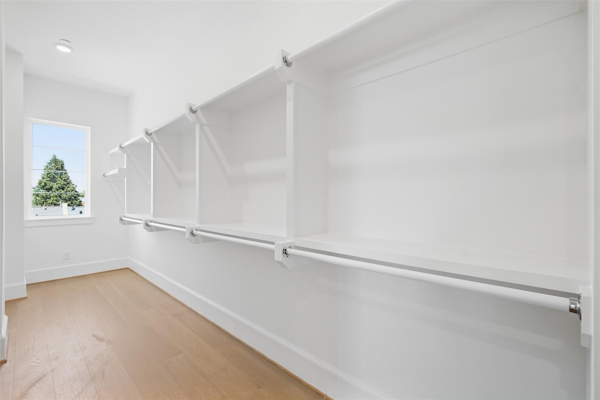Walk-in closet with multiple hanging rods, light wood flooring, and a window overlooking a tree.
