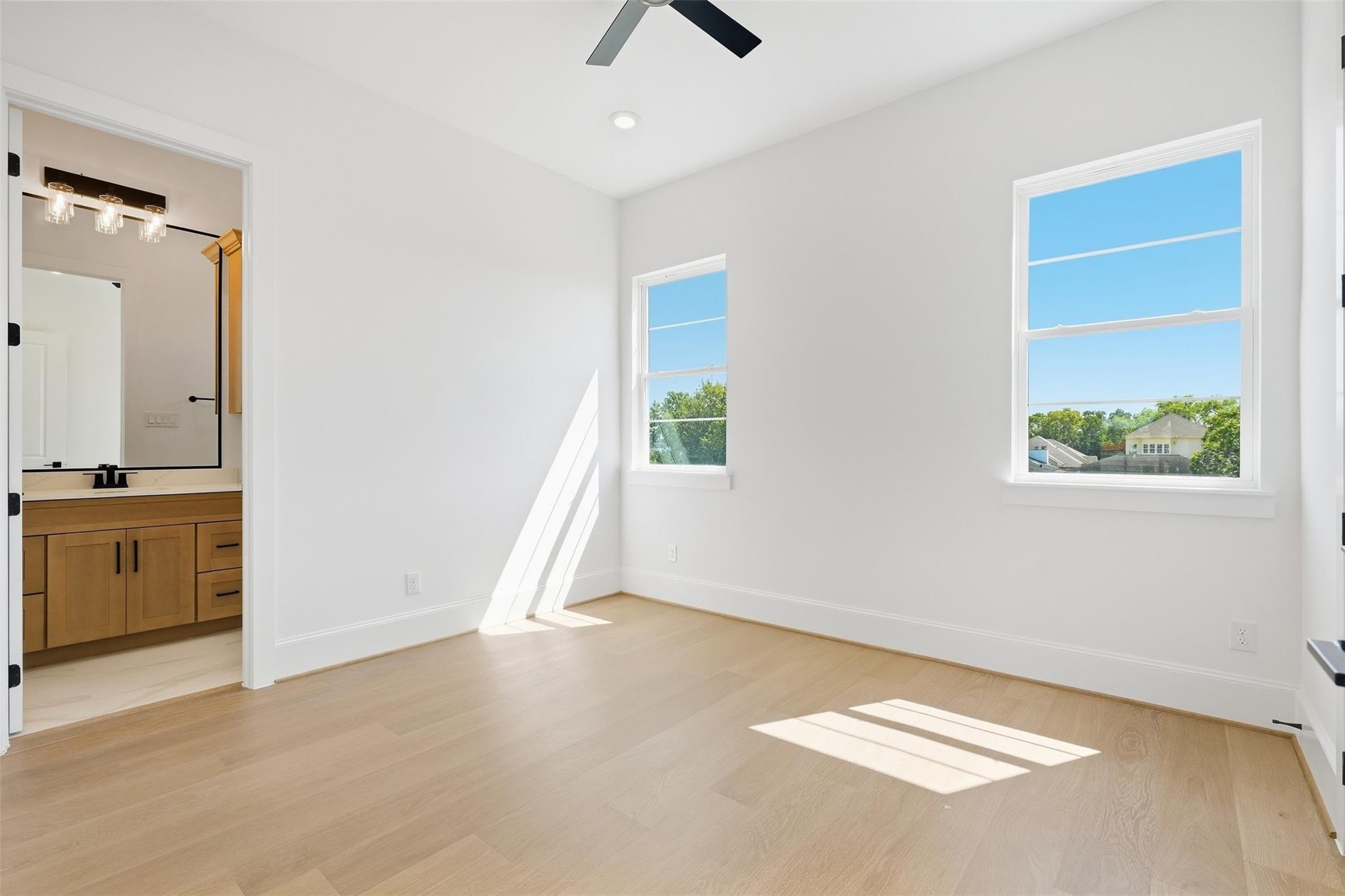 Vacant bedroom with light wood floors, two sunny windows, and an adjoining bathroom vanity.