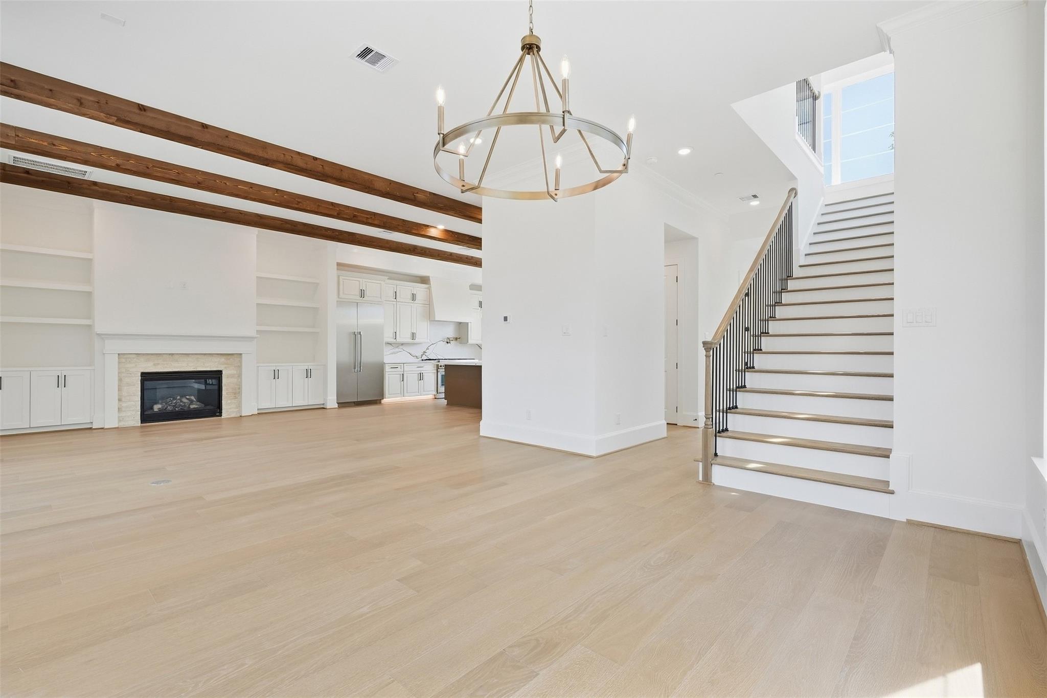 Open living room with exposed wood beams, fireplace, adjoining kitchen, and staircase.