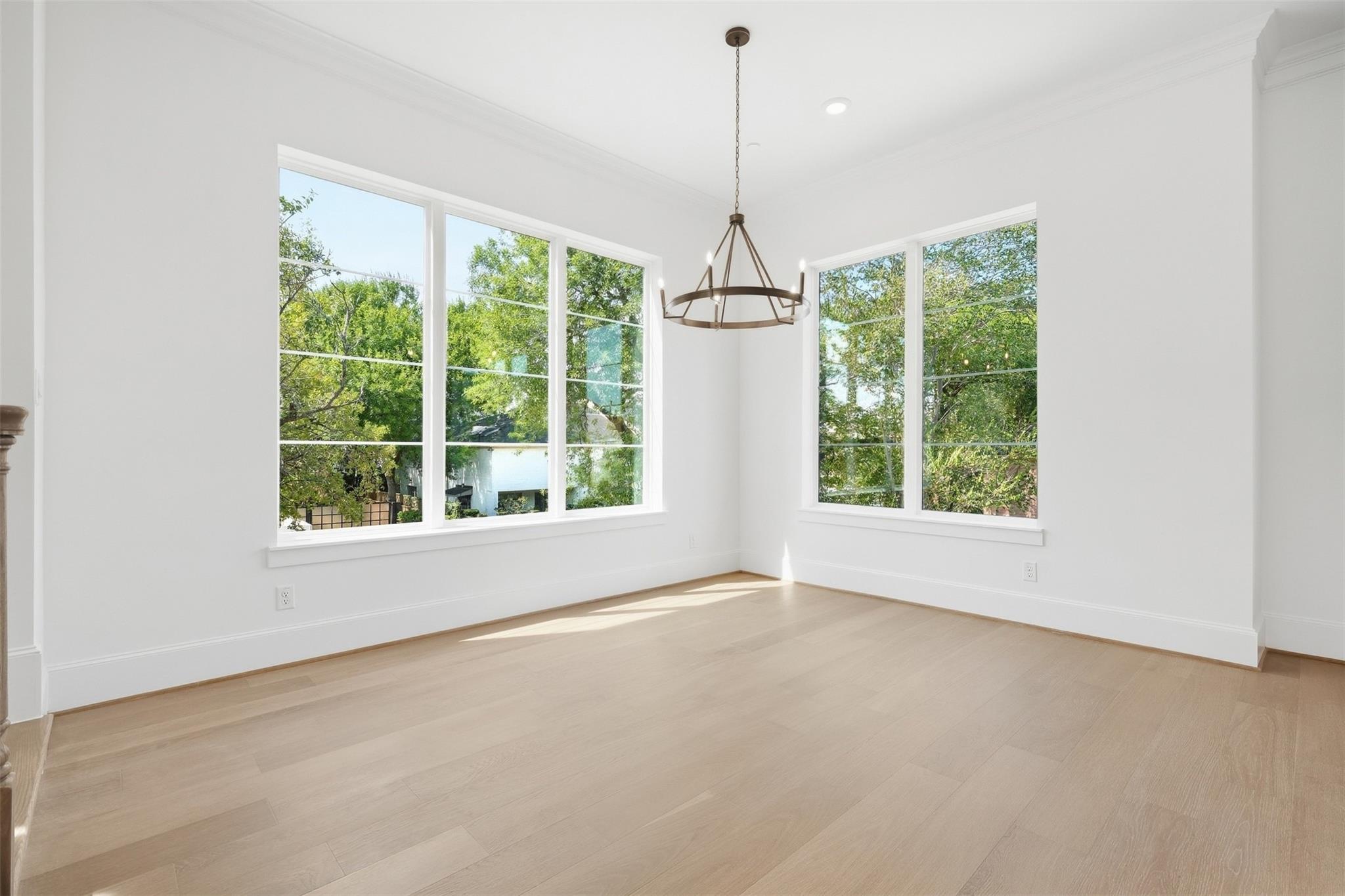 Bright empty room with pale wood floors, a bronze chandelier, and large windows overlooking trees.