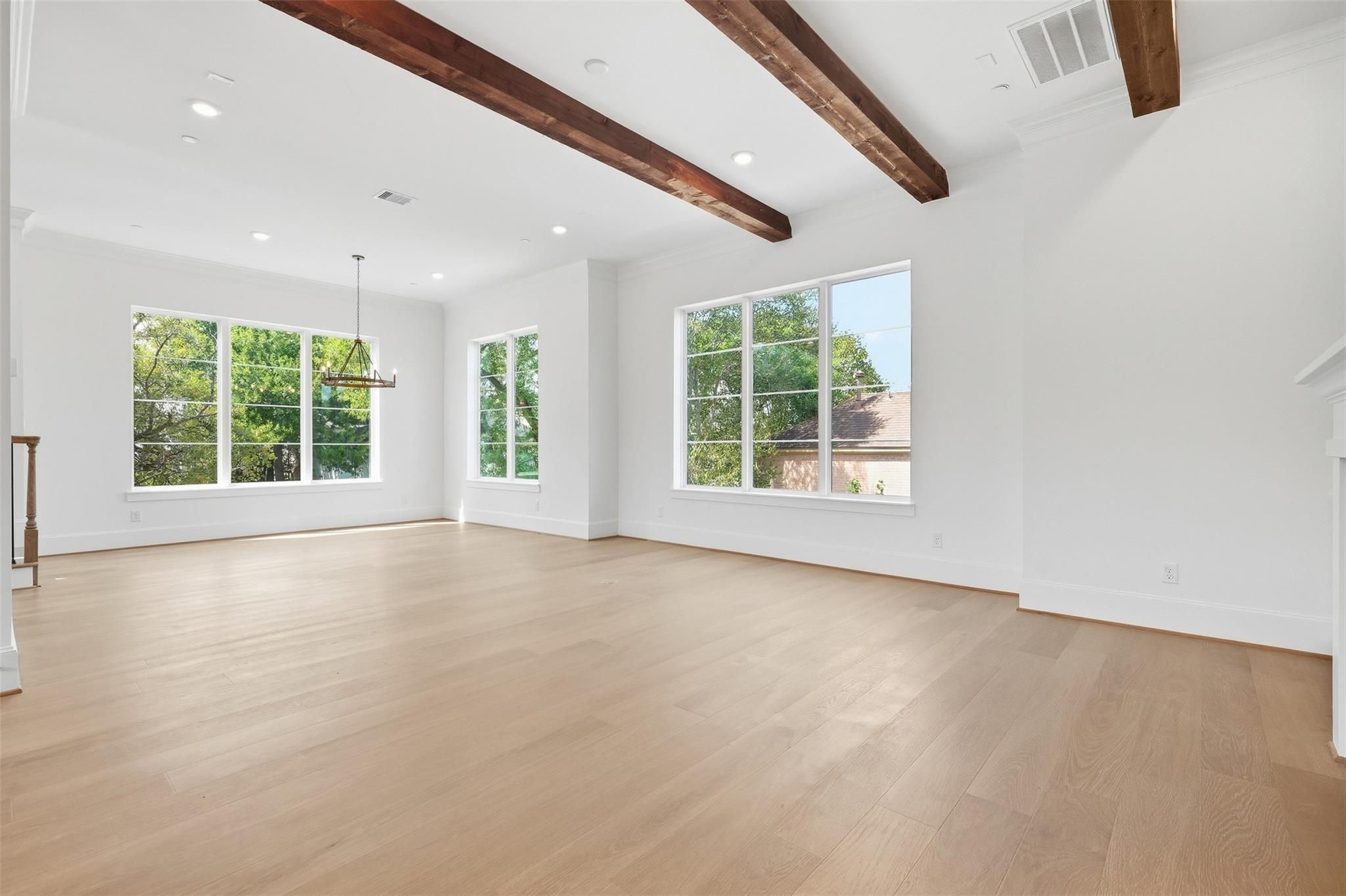 Empty living room with pale wood floors, large windows, and exposed ceiling beams.