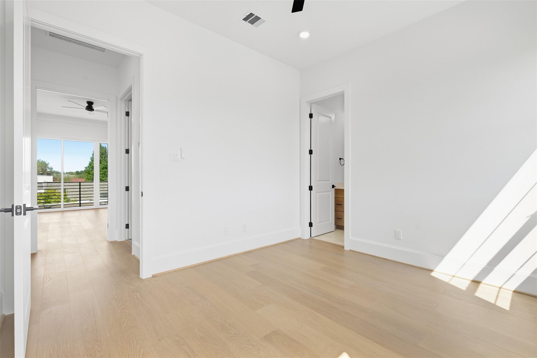 Vacant bedroom with pale wood floors and sunlight, opening to a room with balcony doors.
