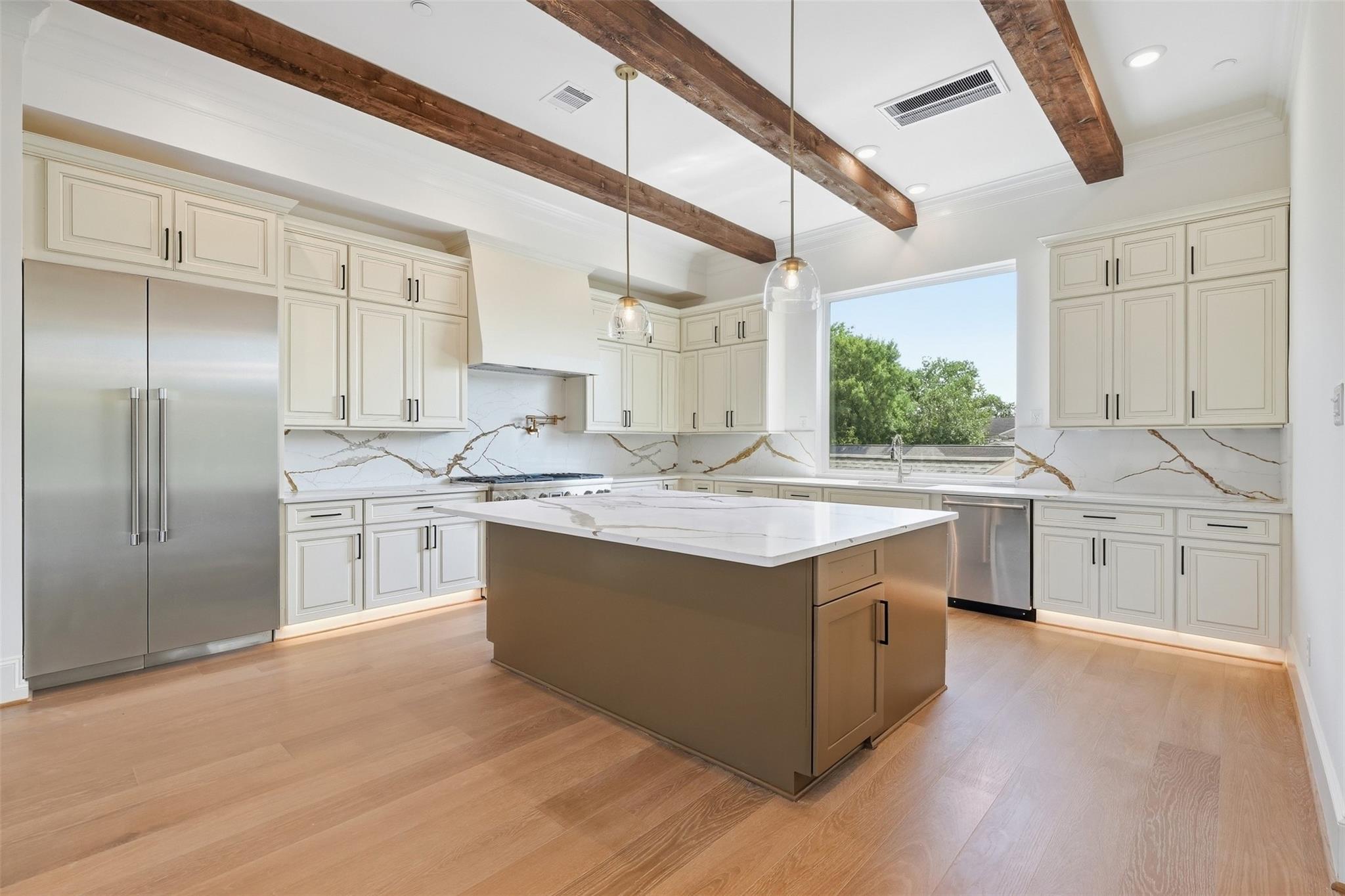 Kitchen with a large island, veined stone backsplash, white cabinets, and exposed wood ceiling beams.