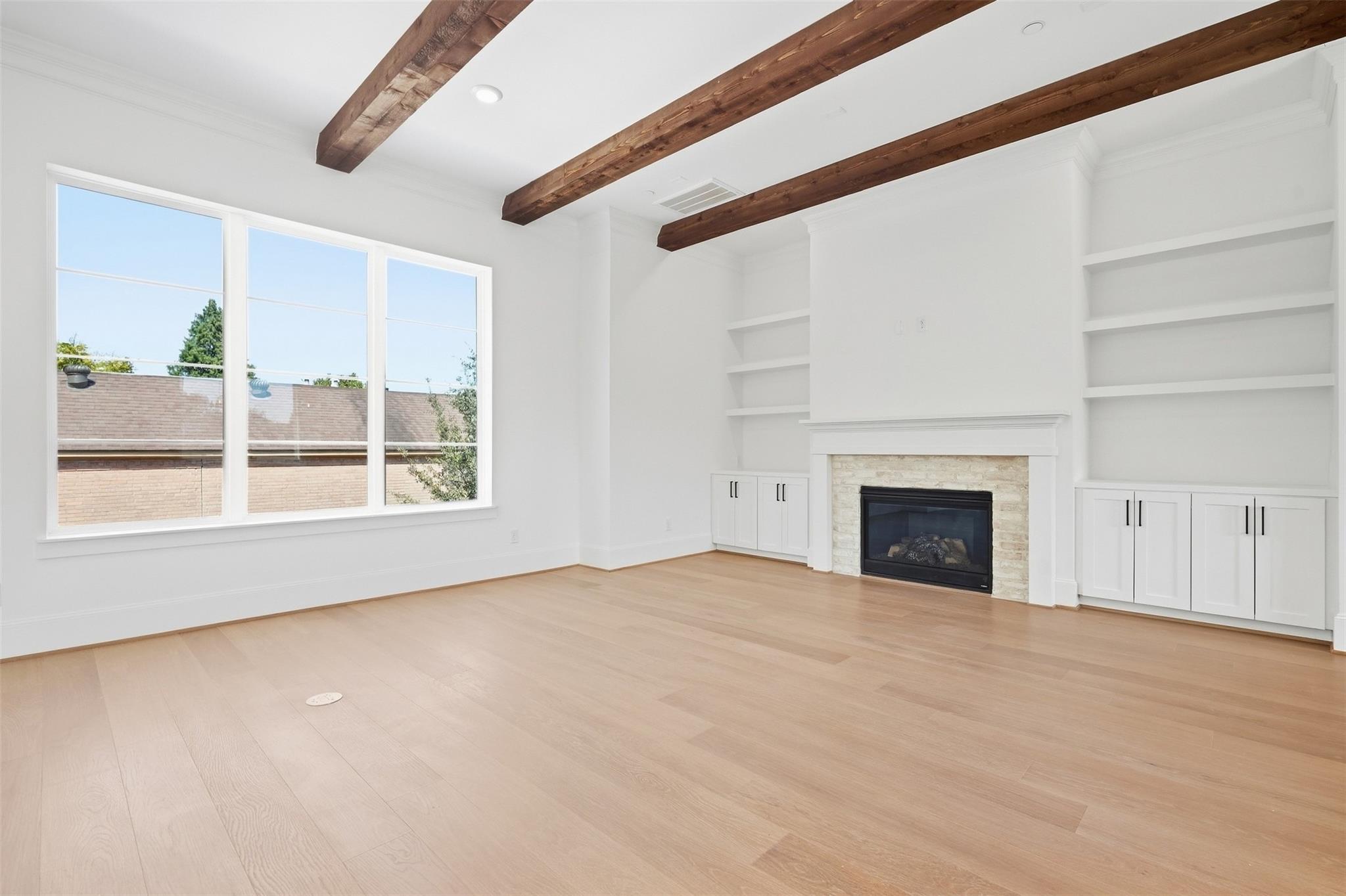 Empty living room with exposed wood ceiling beams, a stone fireplace, built-in shelving, and large windows.