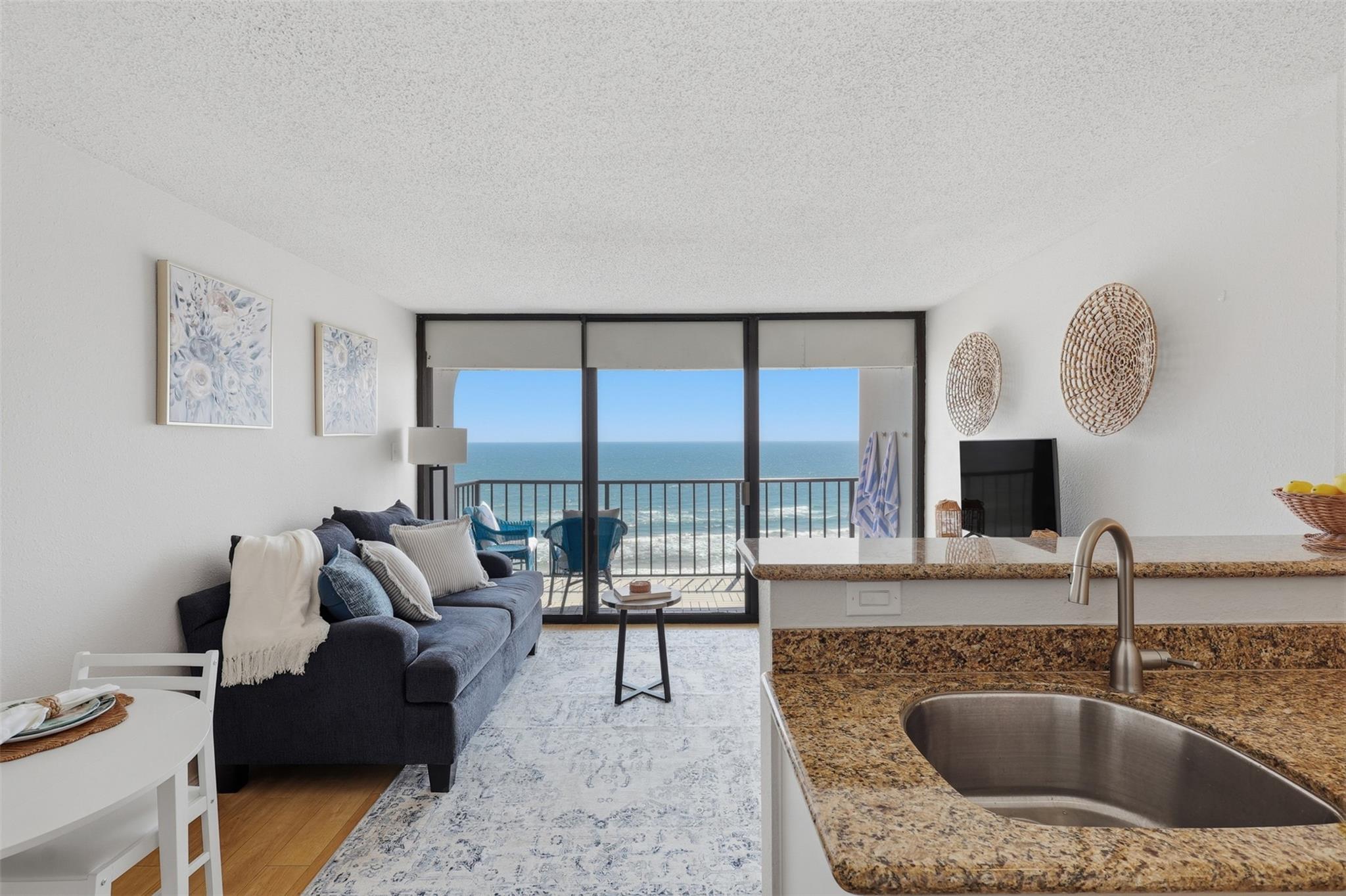 Oceanfront condo living room with a blue sofa, granite kitchen counter, and balcony overlooking the water.
