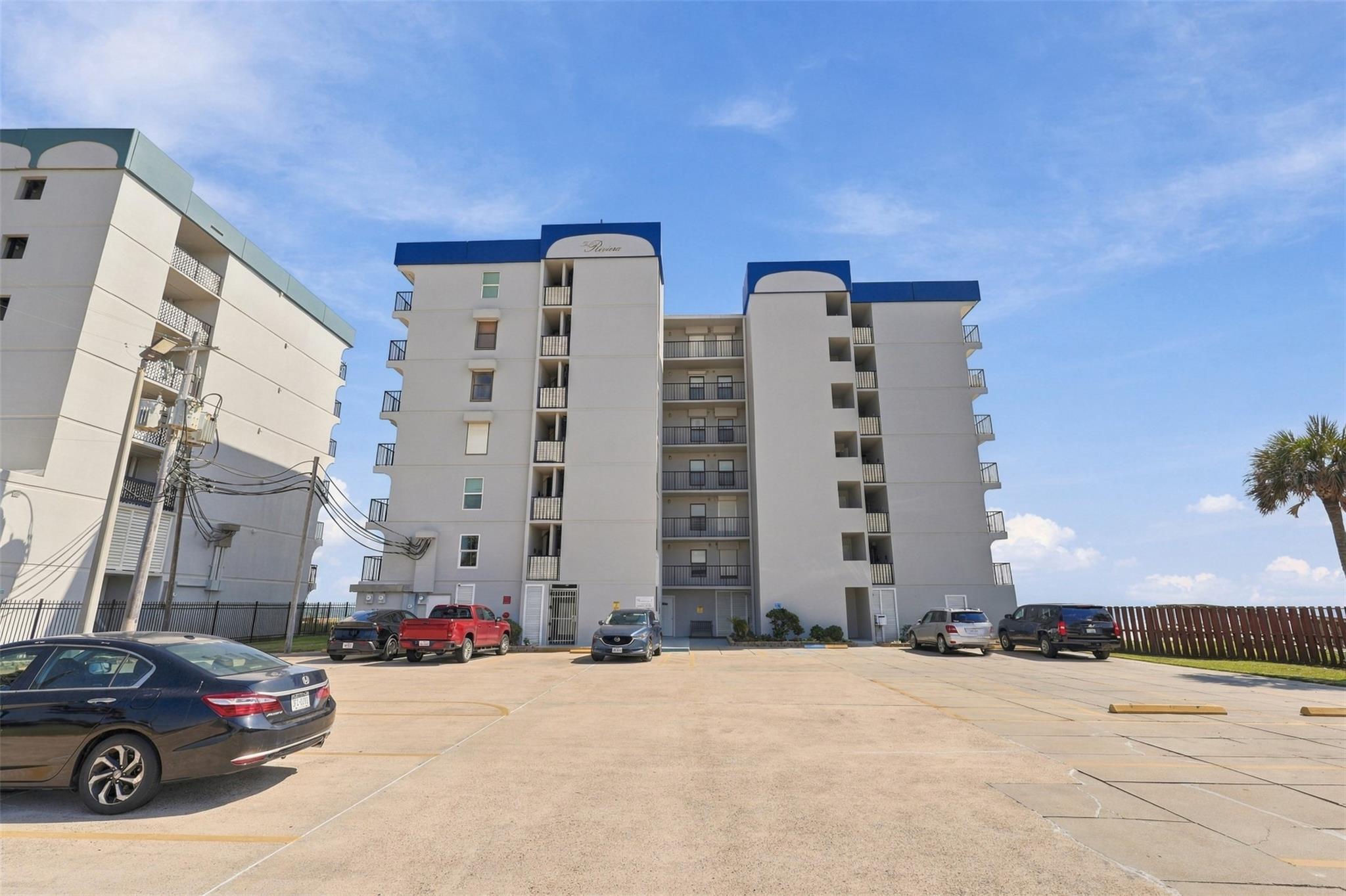 Condominium building with blue roof accents and balconies, viewed from its parking lot.