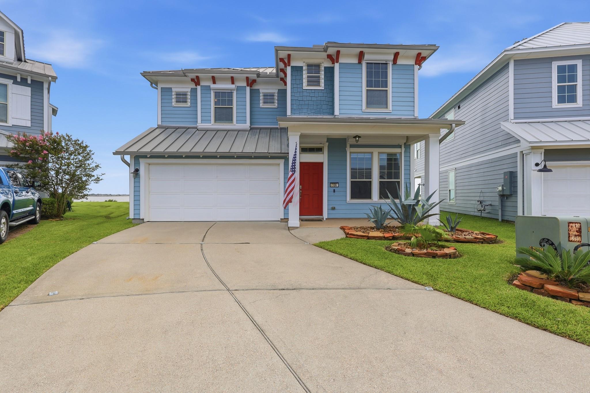 Blue two-story home with a red front door, attached garage, landscaped lawn, and water visible beyond.