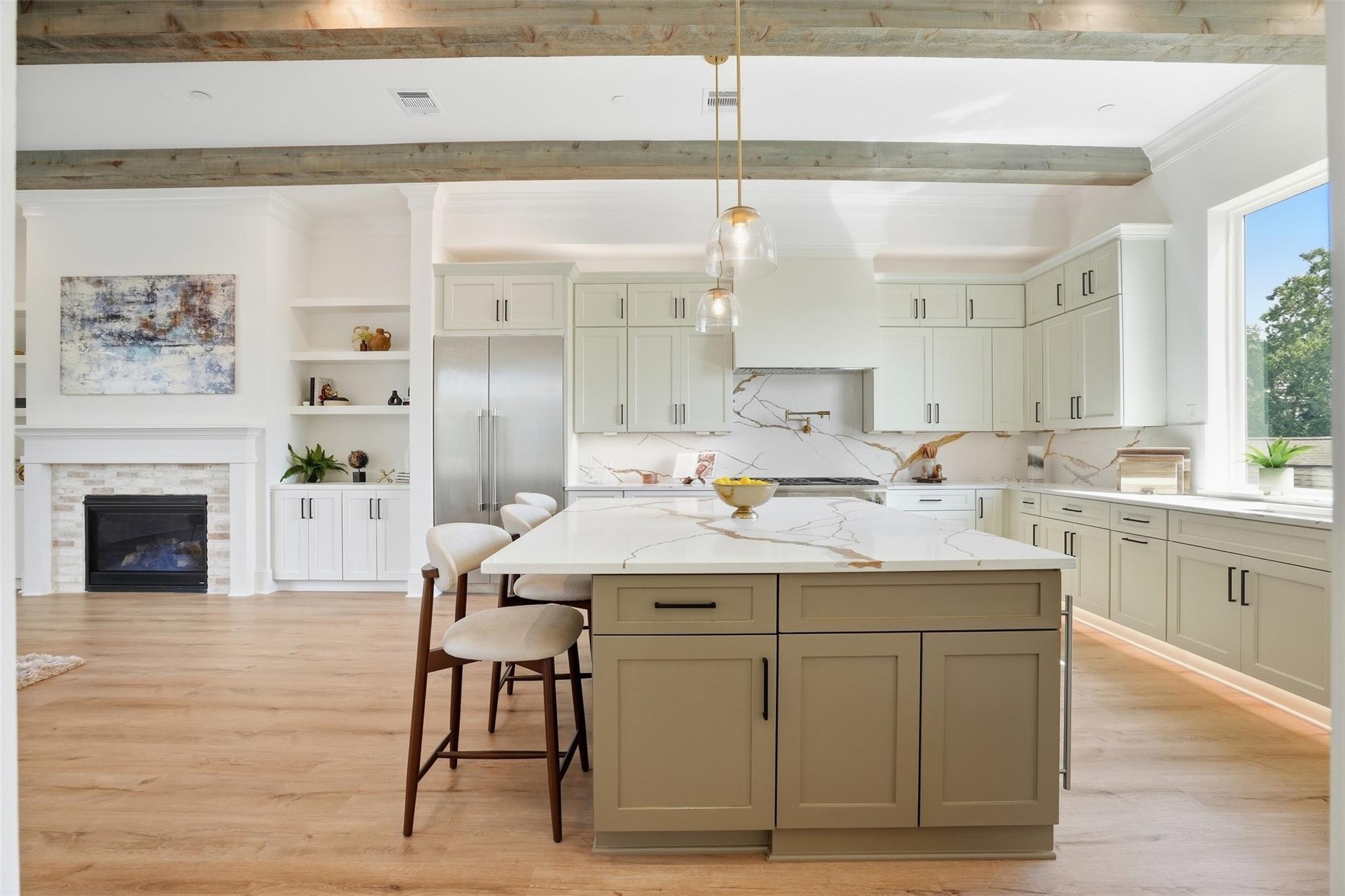 Kitchen with a veined stone island, pale cabinetry, exposed beams, and an adjoining fireplace.