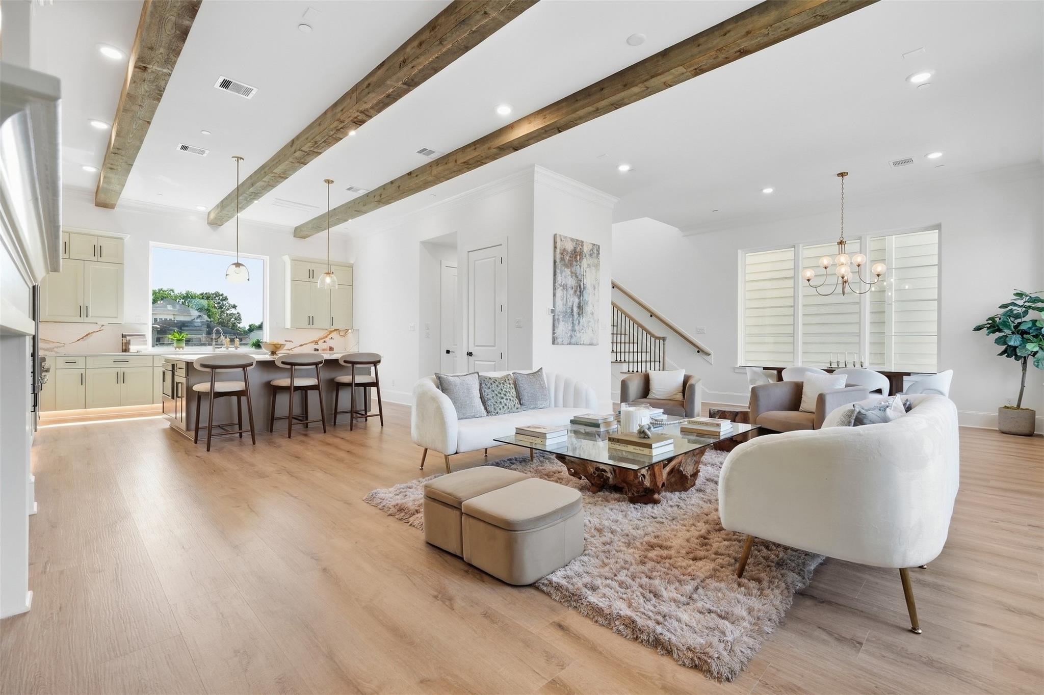 Open-plan living room and kitchen with exposed wood beams, pale floors, and a furnished seating area.