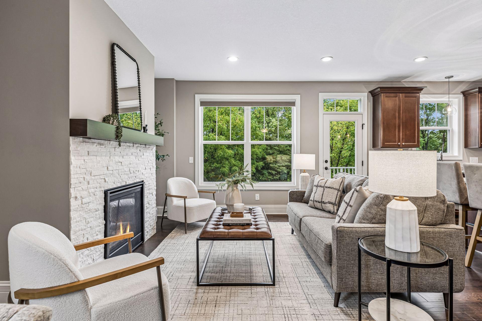 Staged living room with a white stone fireplace, gray sectional, and windows overlooking leafy trees.