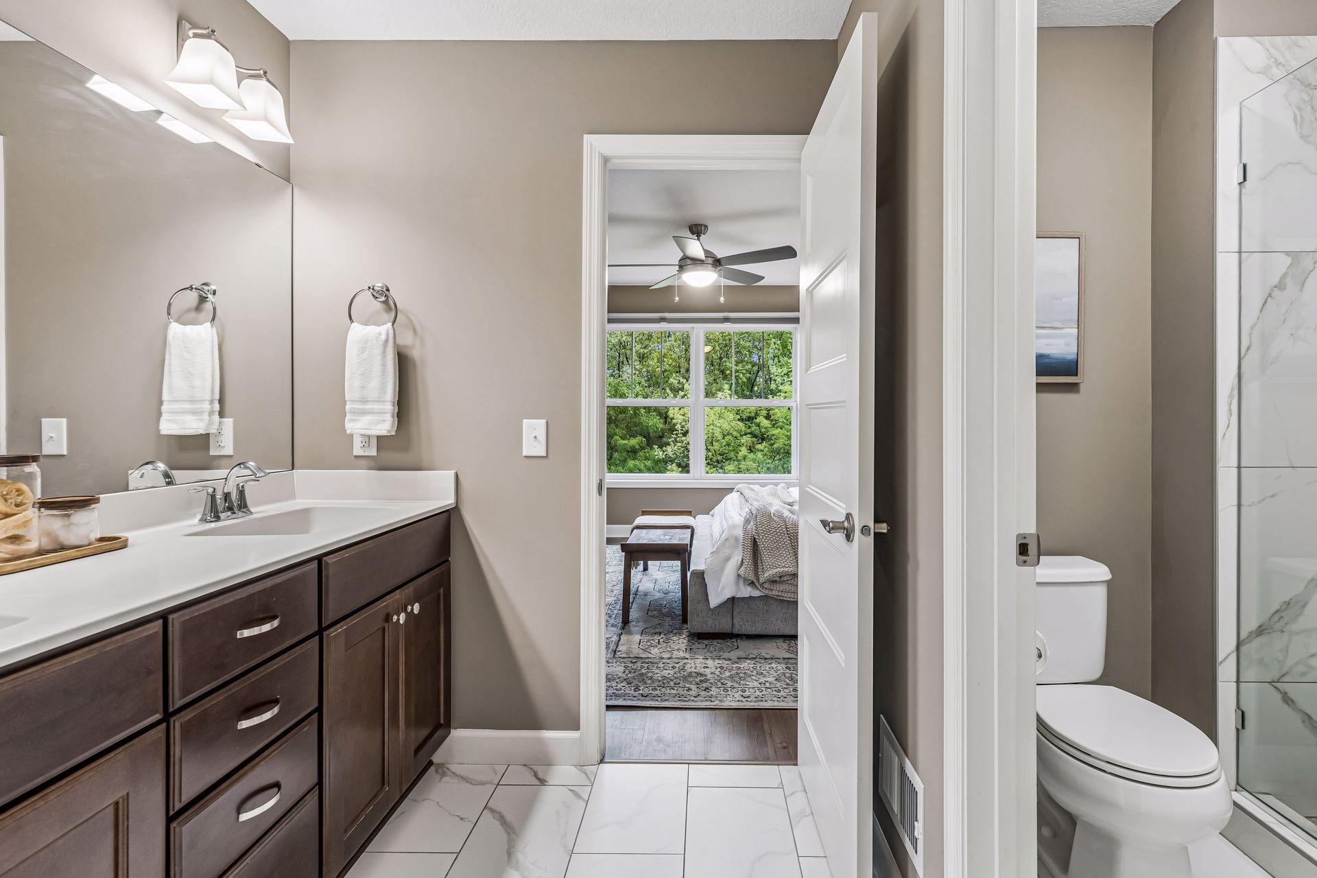 Bathroom with dark wood vanity and marble-look tile, opening onto a bedroom with a leafy window view.