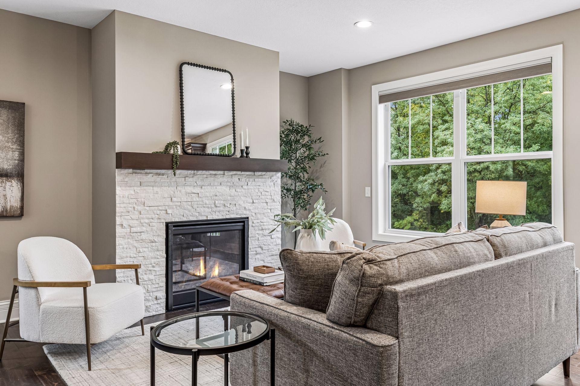 Staged living room with a white stone fireplace and large windows overlooking leafy trees.