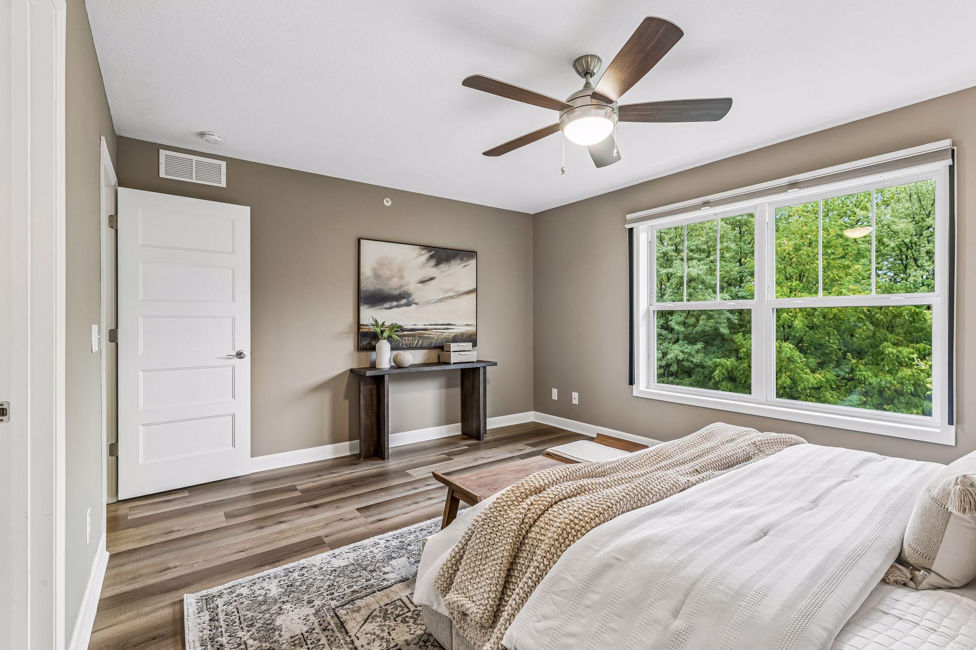 Furnished bedroom with a large window overlooking leafy trees and a wood ceiling fan.