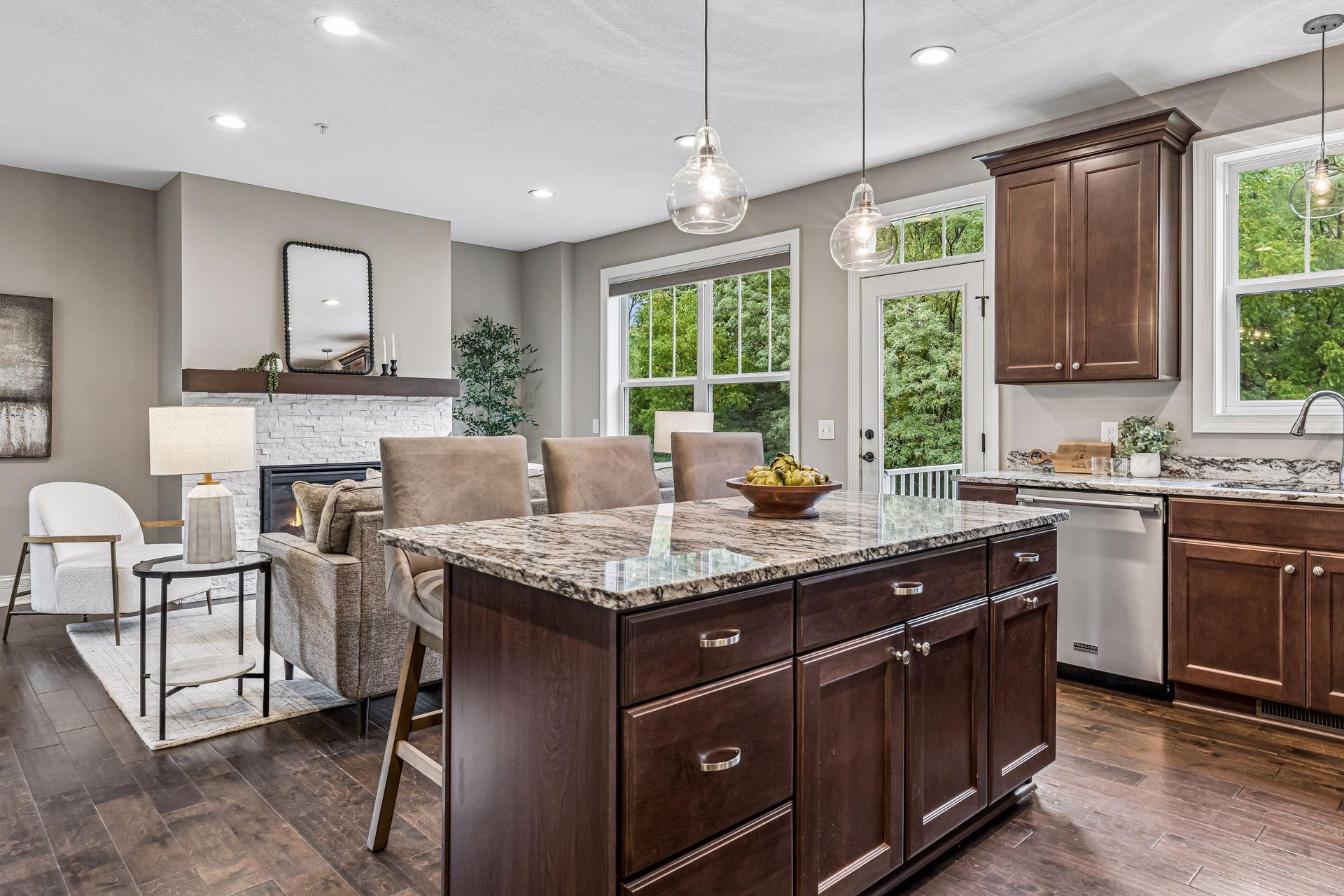 Open-plan kitchen with a granite island, dark wood cabinets, and a white stone fireplace beside leafy windows.