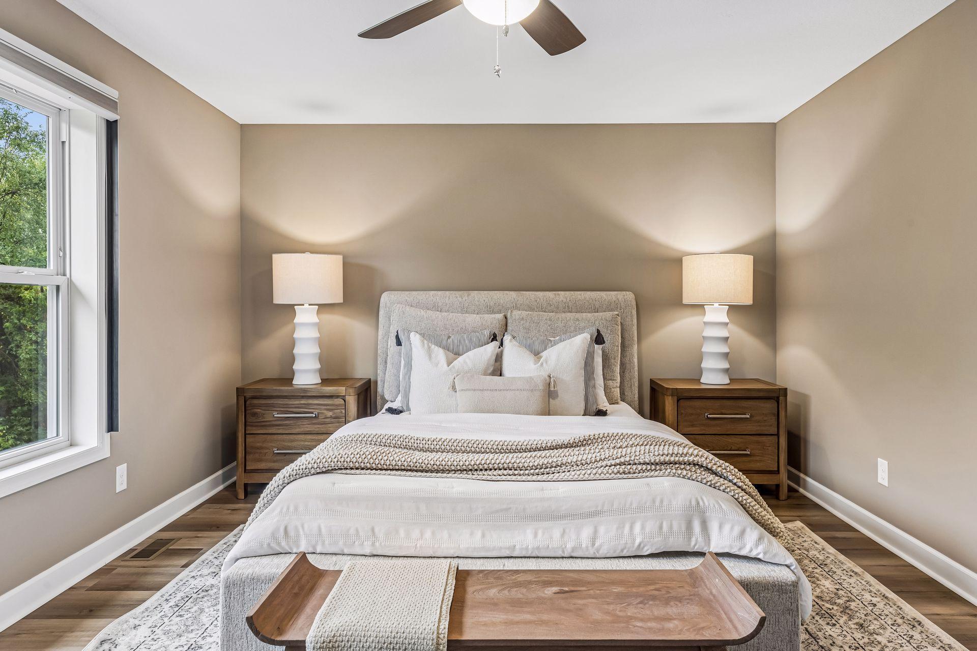 Staged bedroom with a white bed, matching wooden nightstands, and a large window overlooking trees.