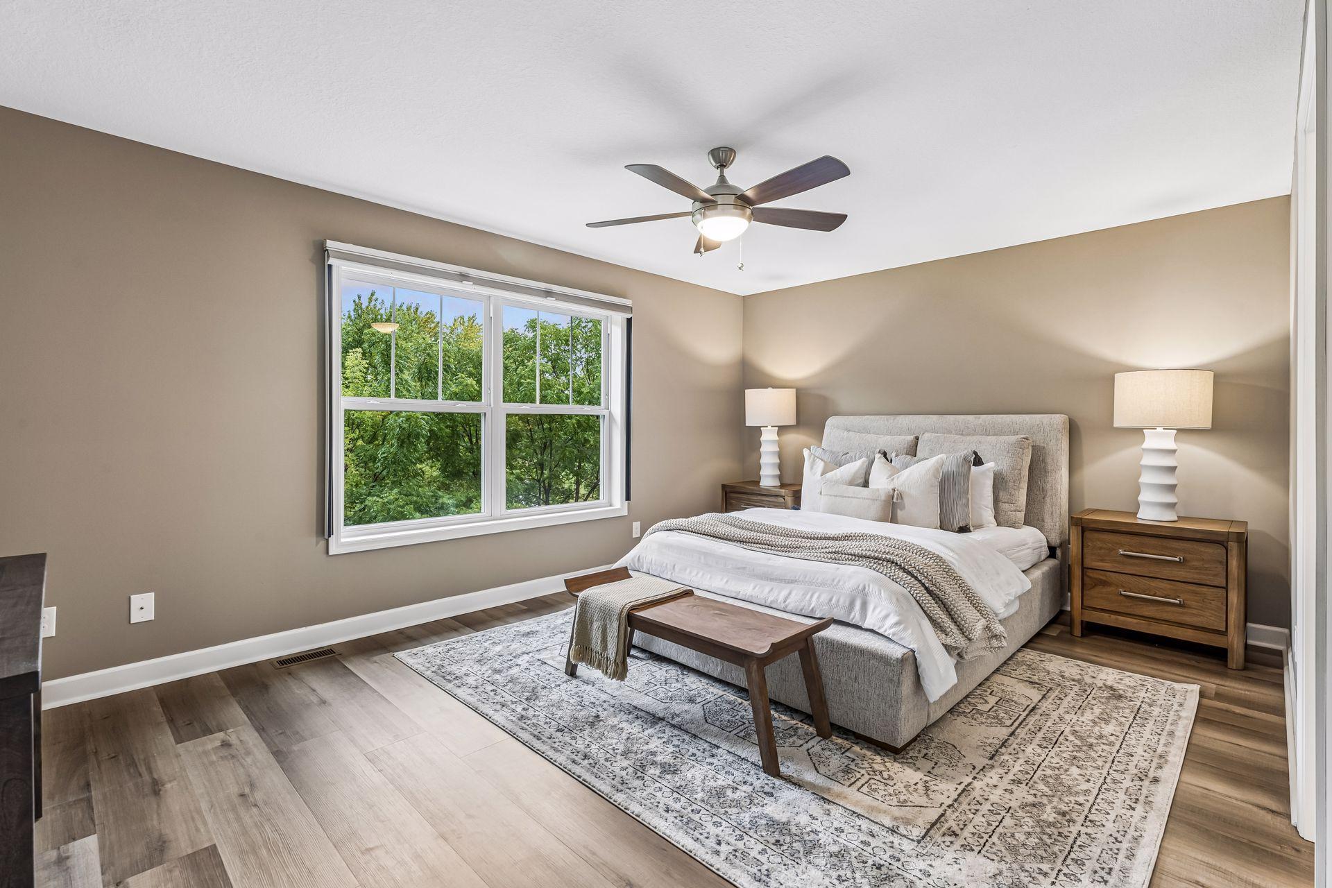 Furnished bedroom with a large window overlooking leafy trees and a ceiling fan above the bed.